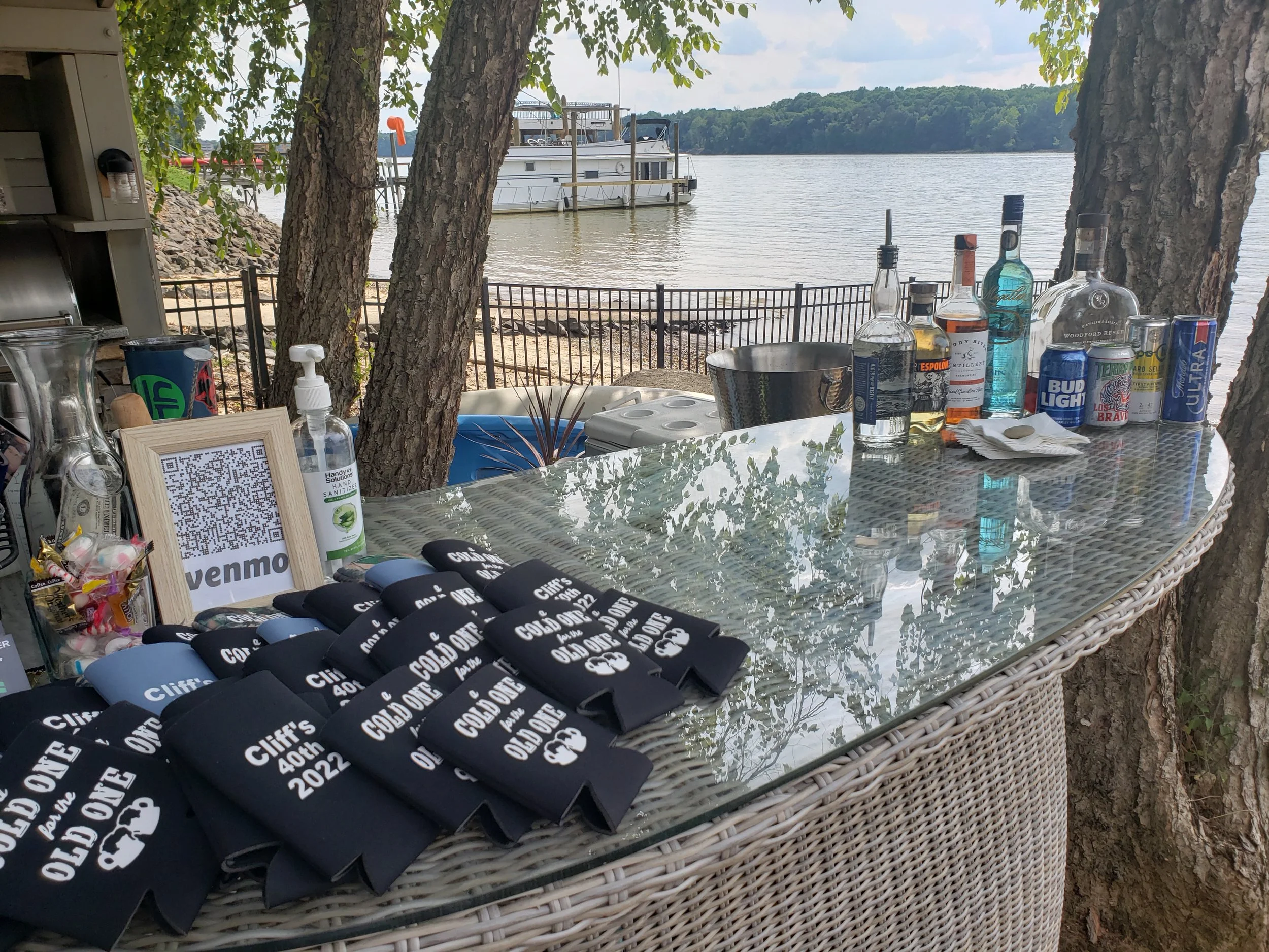 Outdoor party setup by a lakeside with bottles of alcohol, cans of soda, a framed QR code sign, hand sanitizer, and black koozies reading 'Cliff's 40th 2022.'