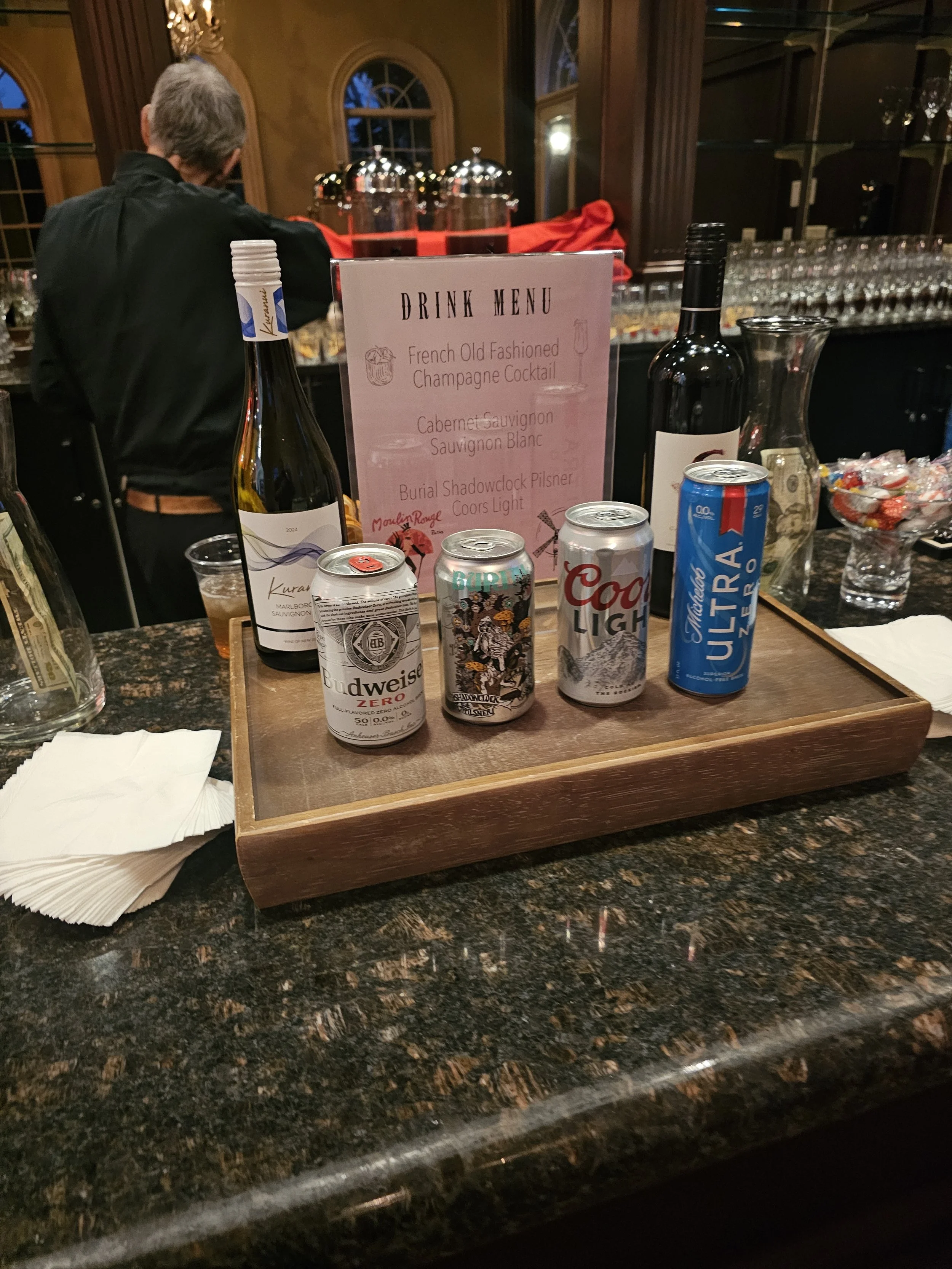 A wooden serving tray on a granite bar counter holding four cans of beer and a wine bottle, with a drink menu sign in the background.