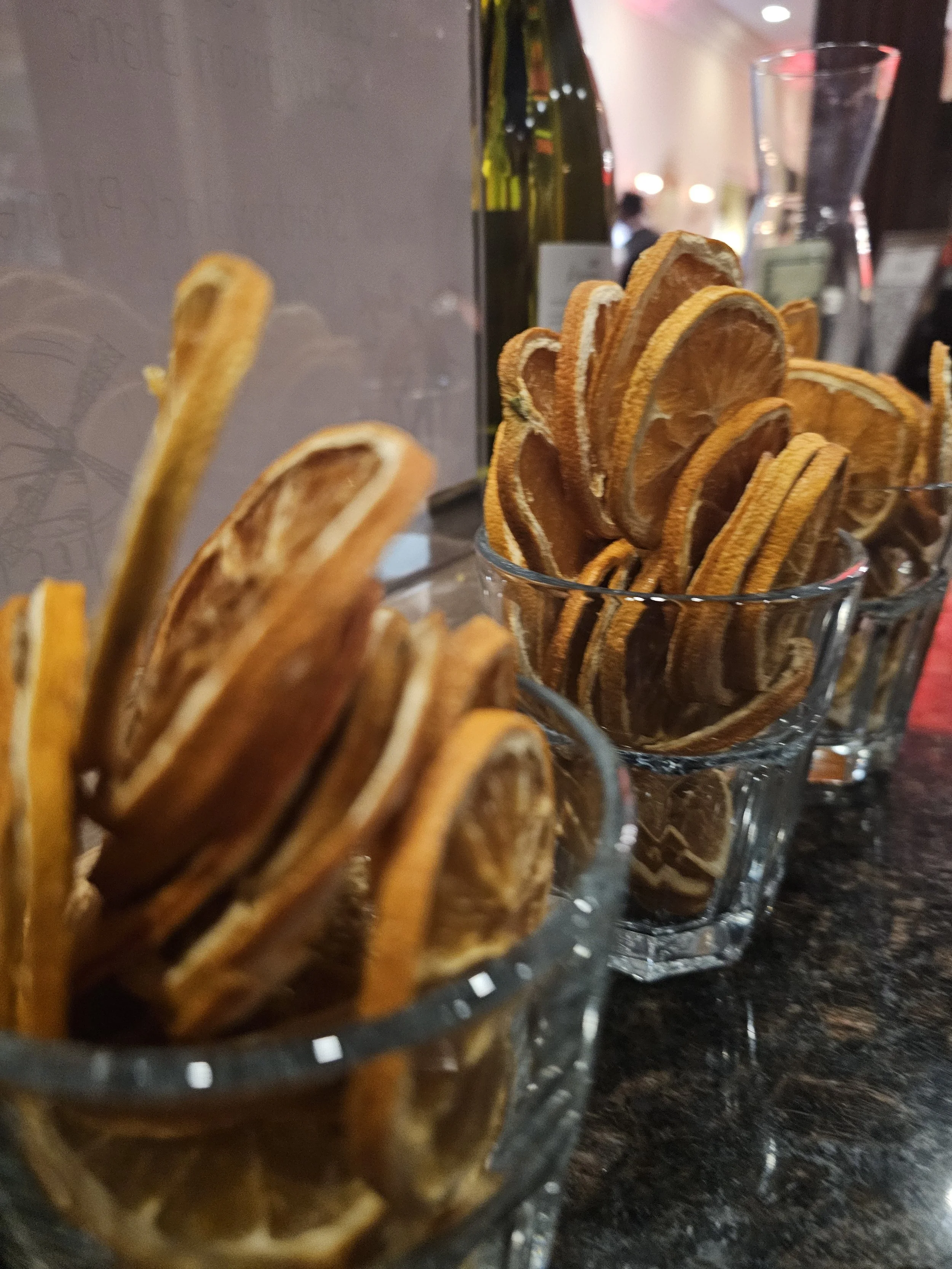 Three glasses filled with dried orange slices on a dark countertop, with bottles and a pitcher in the background.