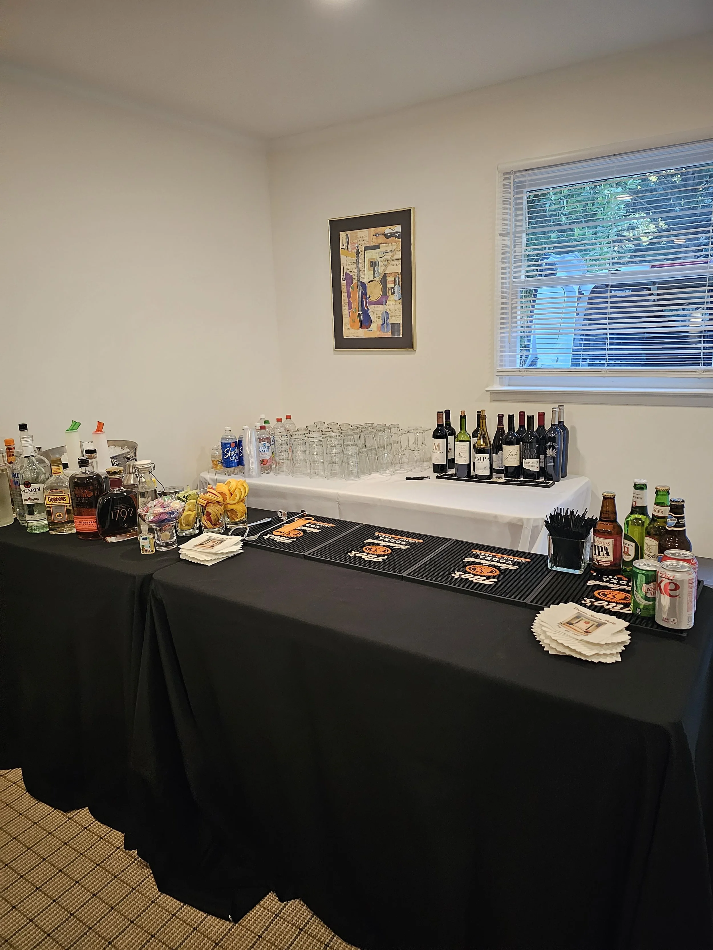 A table set up for serving beverages with various liquor bottles, glasses, snacks, napkins, and soda cans inside a room with a window and wall art.