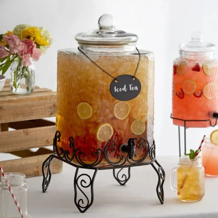 Large glass jar with lemon-infused iced tea on a black metal stand, with a chalkboard tag reading 'Iced Tea'; in the background, smaller jars of fruit-infused drinks and a wooden crate with flowers.
