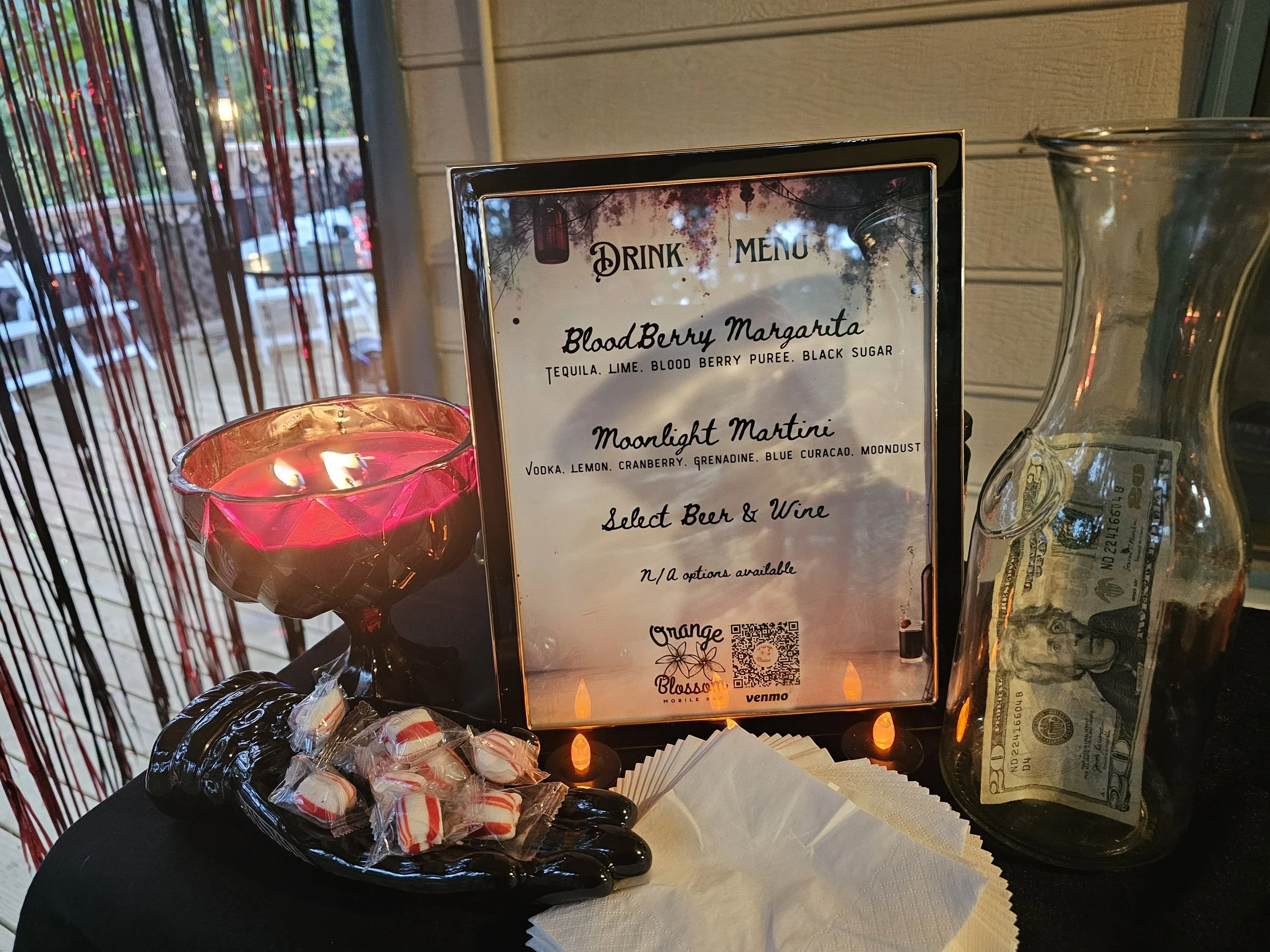 Table with a lit red candle in a glass holder, a black hand-shaped dish with small wrapped candies, a menu sign listing drinks including Blood Berry Margarita and Moonlight Martini, and a jar containing several U.S. dollar bills. The background featu
