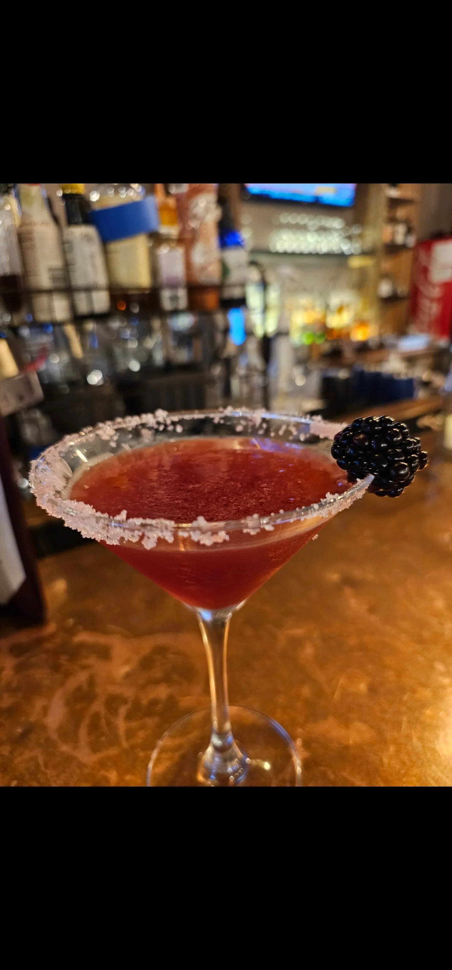 Red cocktail in a martini glass garnished with a blackberry and salt rim, set on a bar counter.
