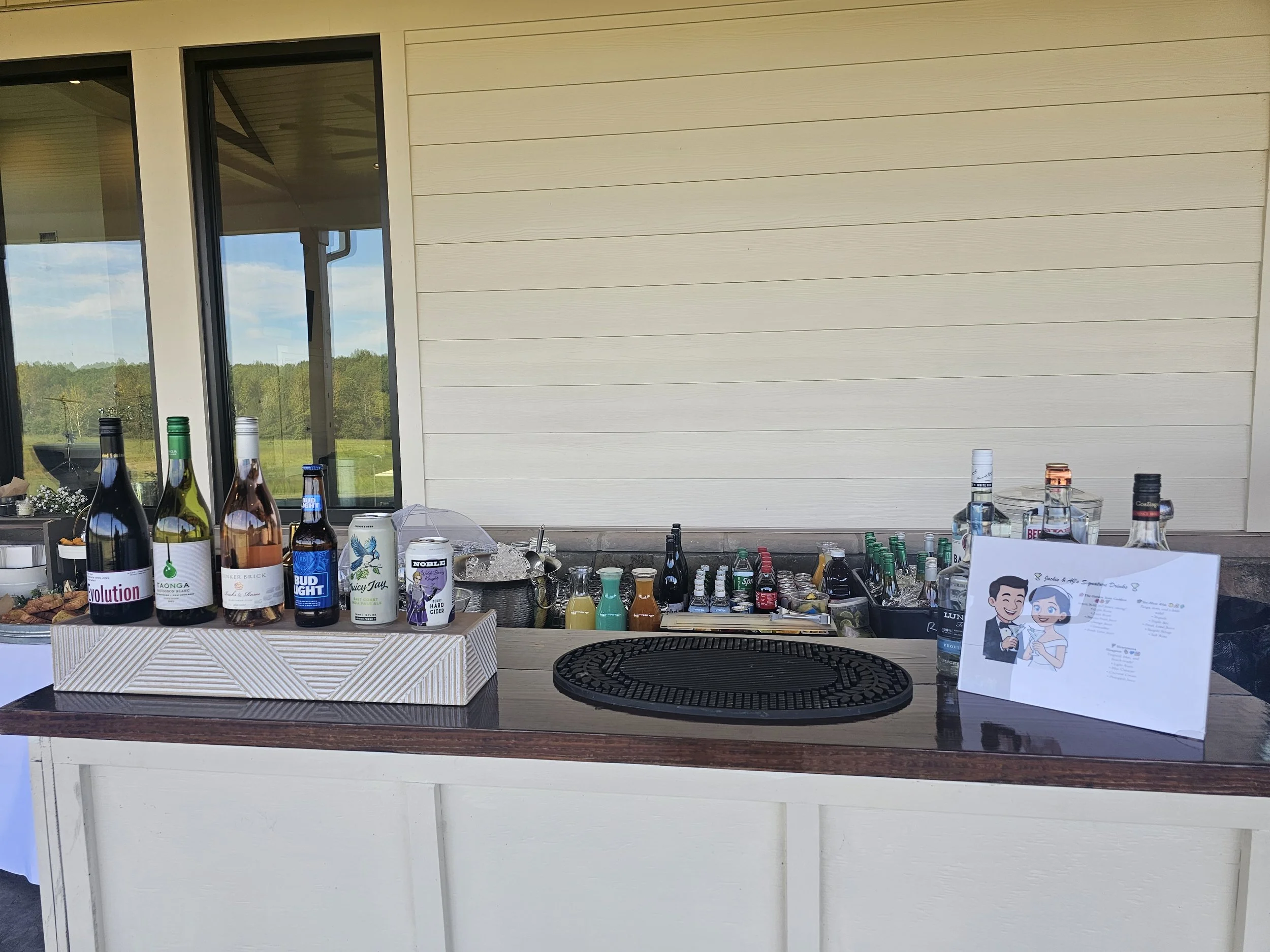 Outdoor bar setup with liquor bottles, mixers, and a wedding or event sign, with windows showing a green outdoor landscape.