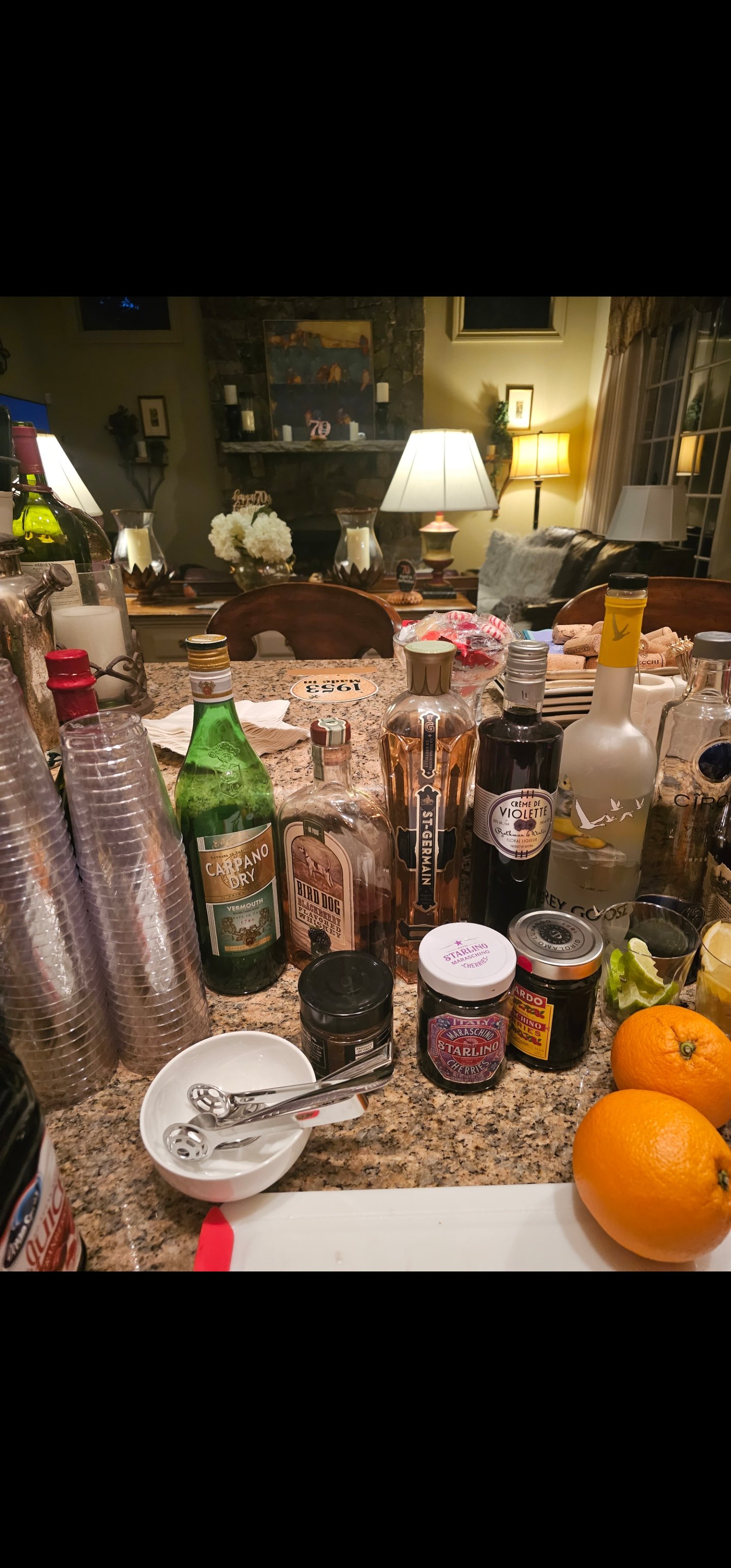 A cluttered kitchen counter with various bottles, jars, oranges, a white bowl, and bar tools in a home setting.