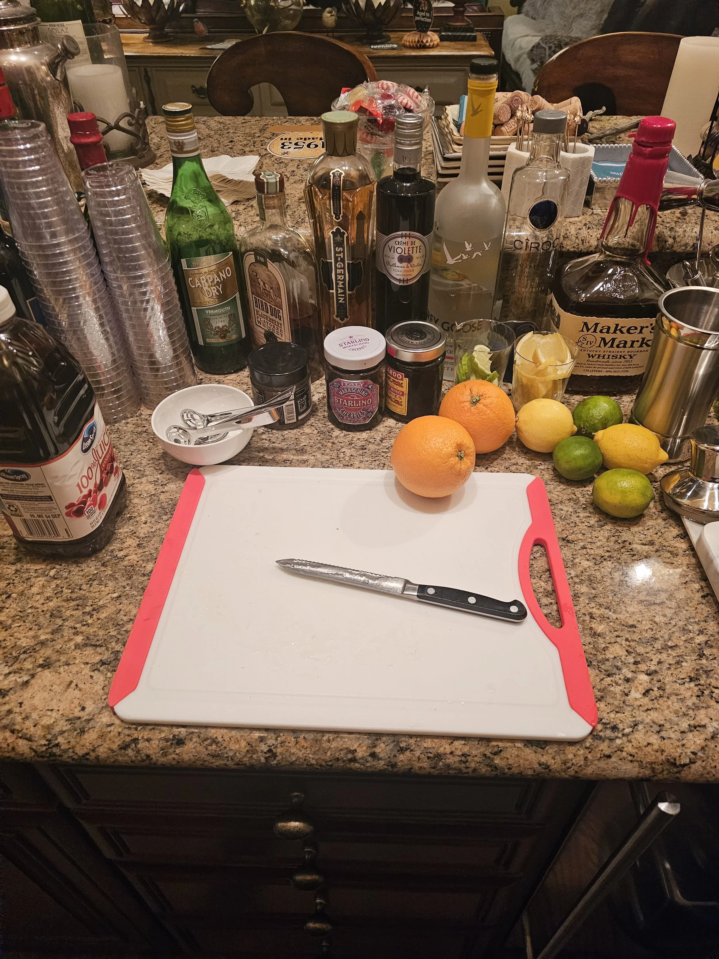 Kitchen counter with lemons, limes, a cutting board, a knife, various liquor bottles, jars, and cups arranged for cocktail preparation.