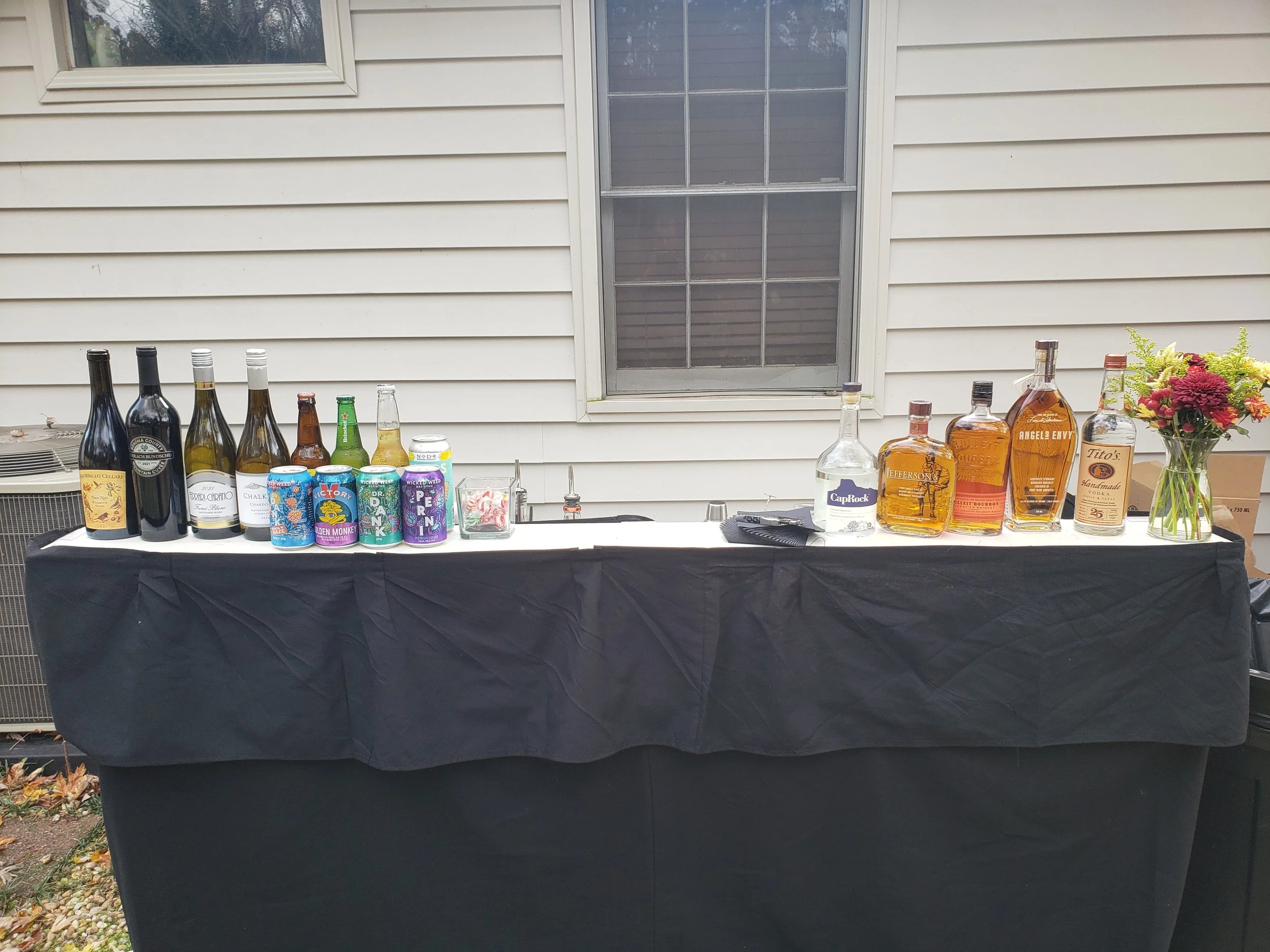 A table with various bottles of alcohol, cans, and a flower arrangement in a glass jar against the exterior wall of a house.