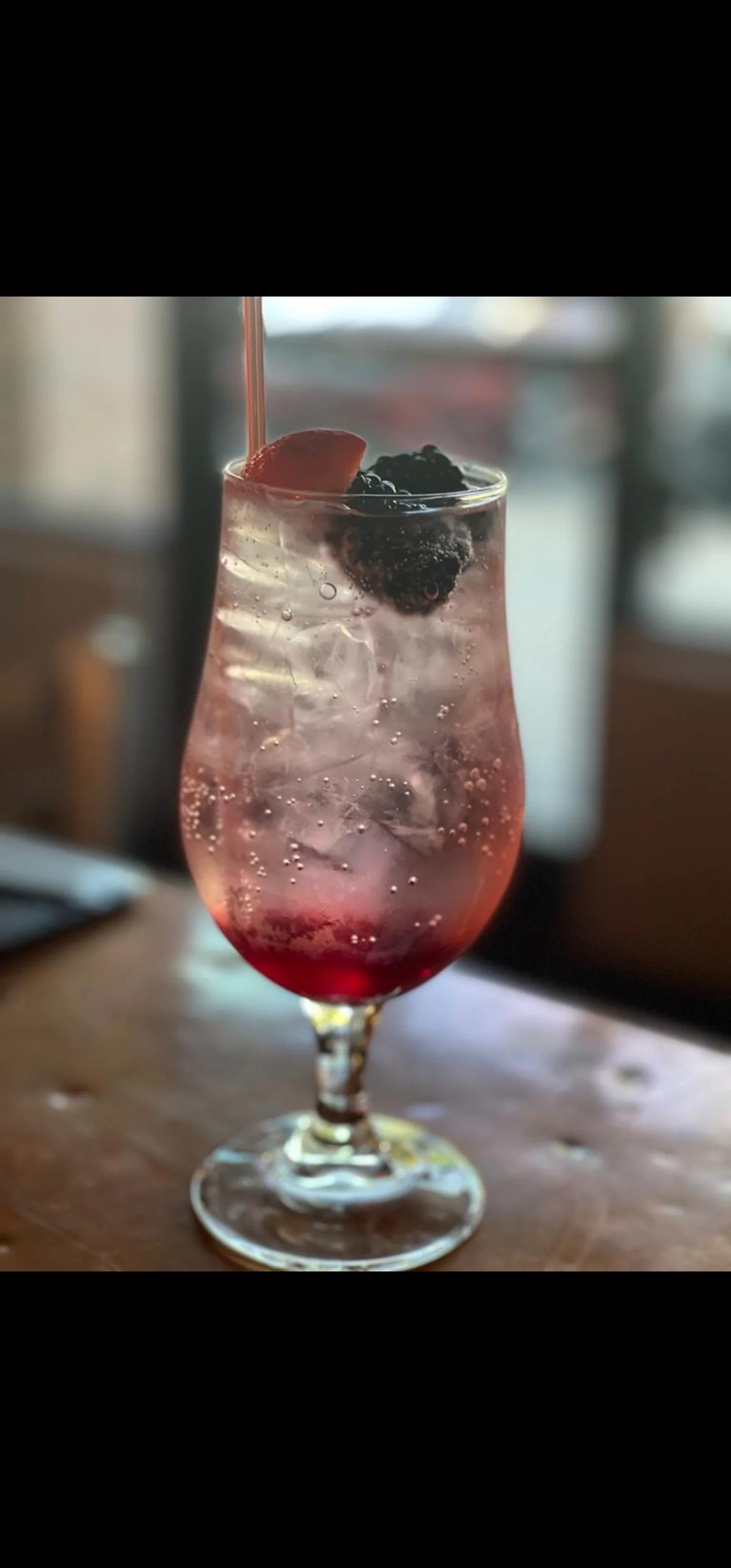 A tall, curvy glass of pink and clear soda with ice, blackberries, and a strawberry on top, sitting on a wooden table in a cozy indoor setting.