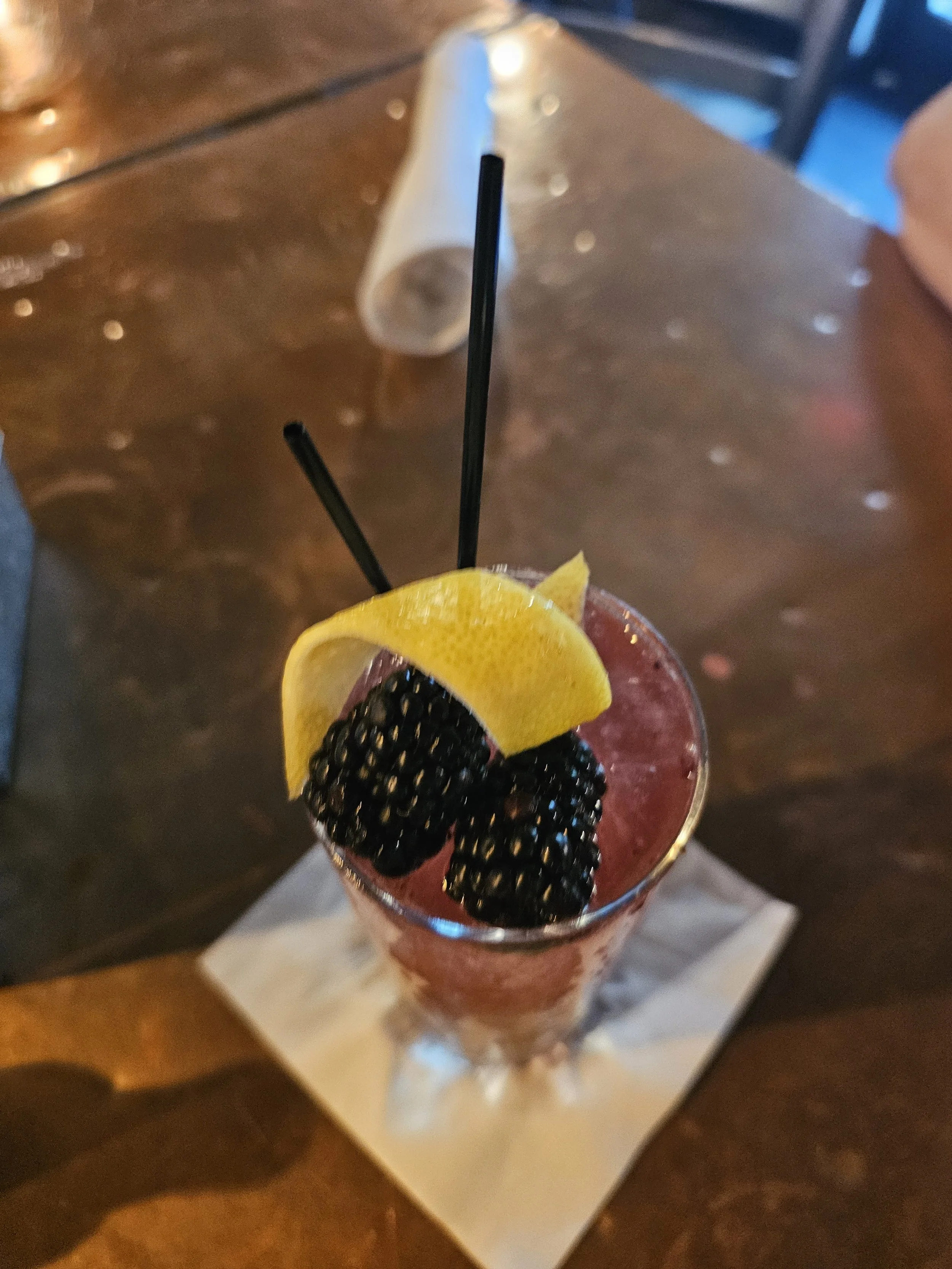 A tall glass of berry cocktail garnished with lemon wedge, blackberries, and two black straws, placed on a napkin on a wooden table.