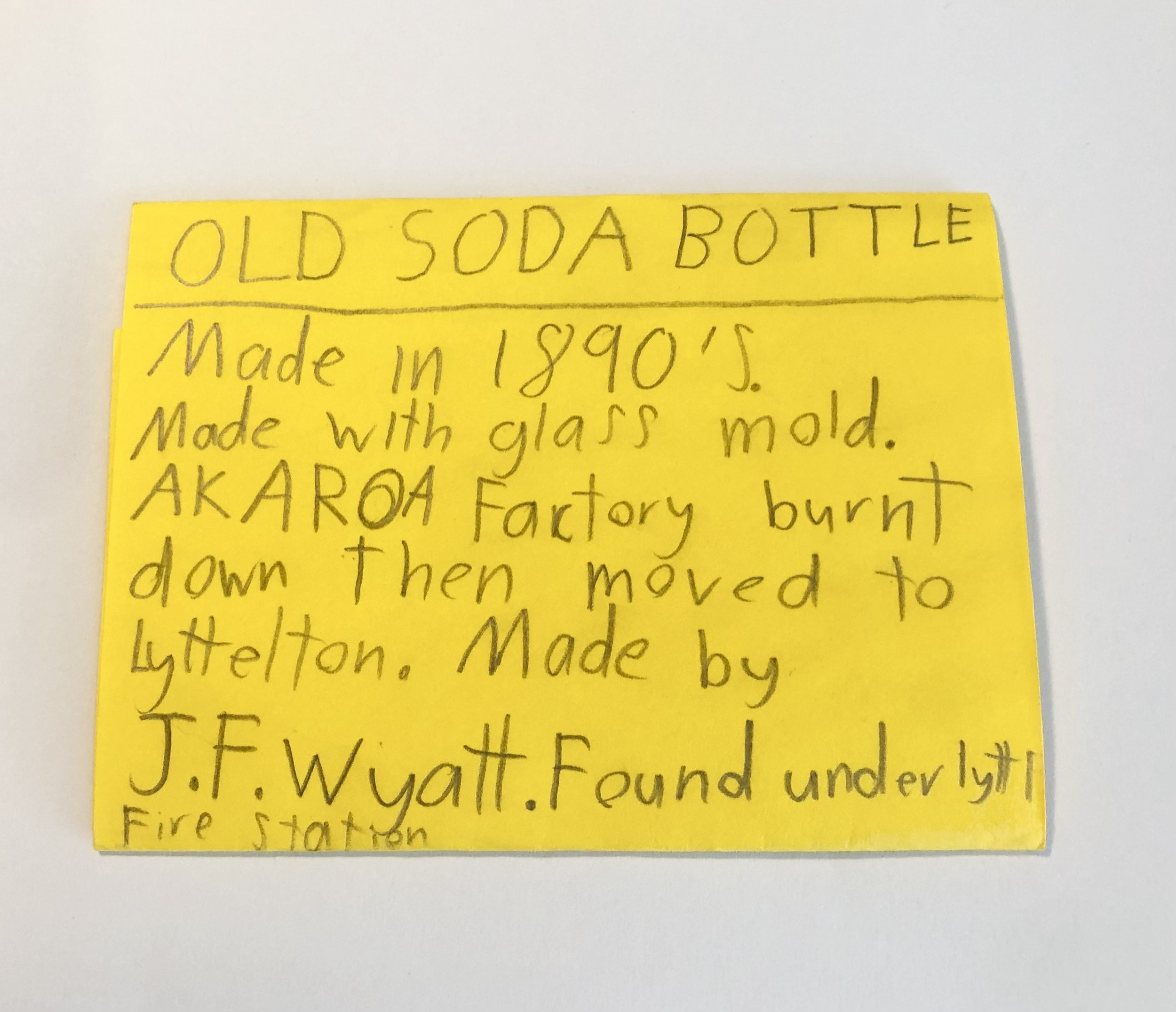 Handwritten note on yellow paper about an old soda bottle, mentioning it was made in 1890s with glass mold at AKAROA Factory, burnt, then moved to Lyttelton, by J.F. Wyatt under the Lyttelton Fire Station.