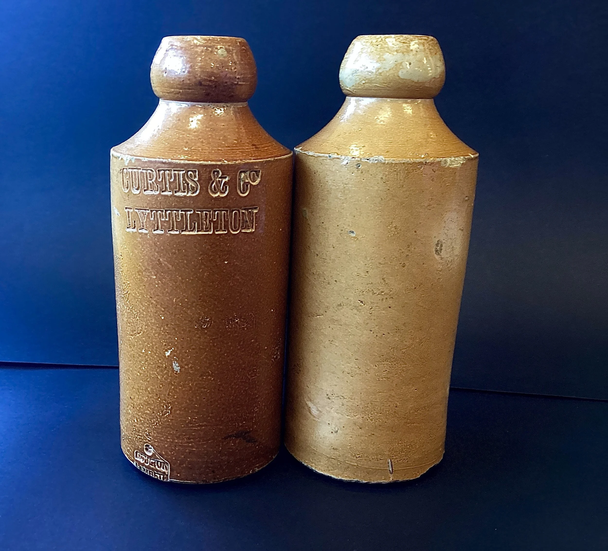 Two vintage ceramic bottles with cork stoppers, one labeled 'Curtis & C. Littleton' in raised lettering, against a dark background.