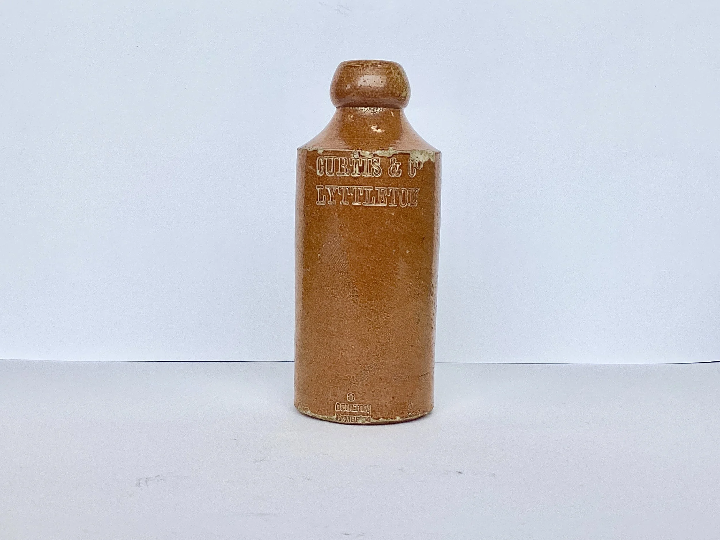 A vintage brown ceramic bottle with embossed text that reads 'CURTIS & CO LYTTELTON.' The bottle is upright on a plain surface with a plain white background.