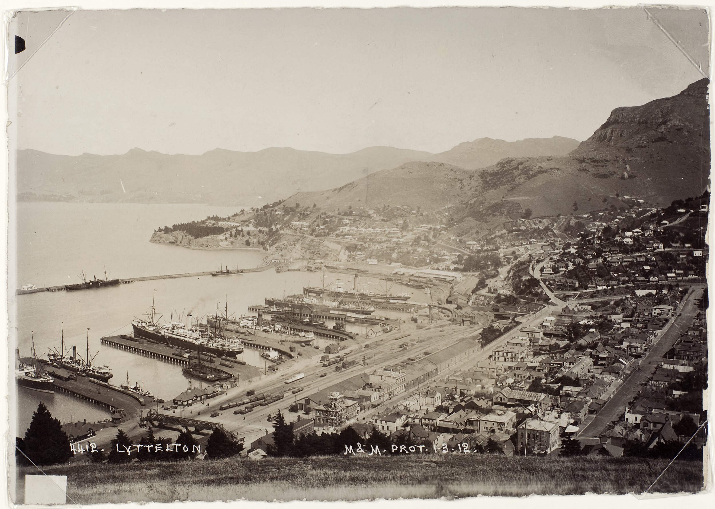 Historical black and white photograph of the port town of Lyttelton, showing ships docked at the harbor, buildings along the waterfront, and hills in the background.