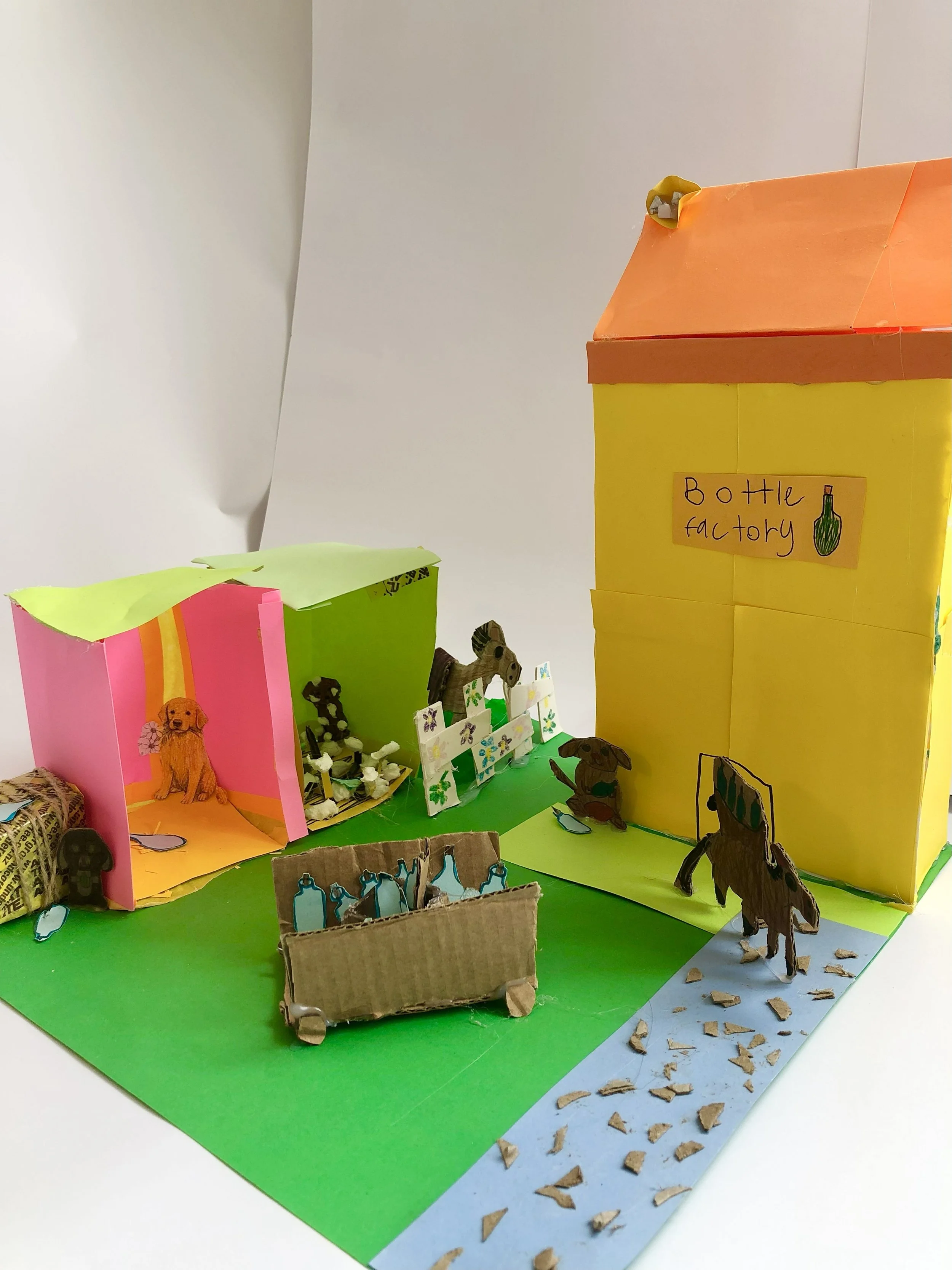 A colorful diorama of a bottle factory with paper animals and bottles, made from paper, cardboard, and other craft materials.