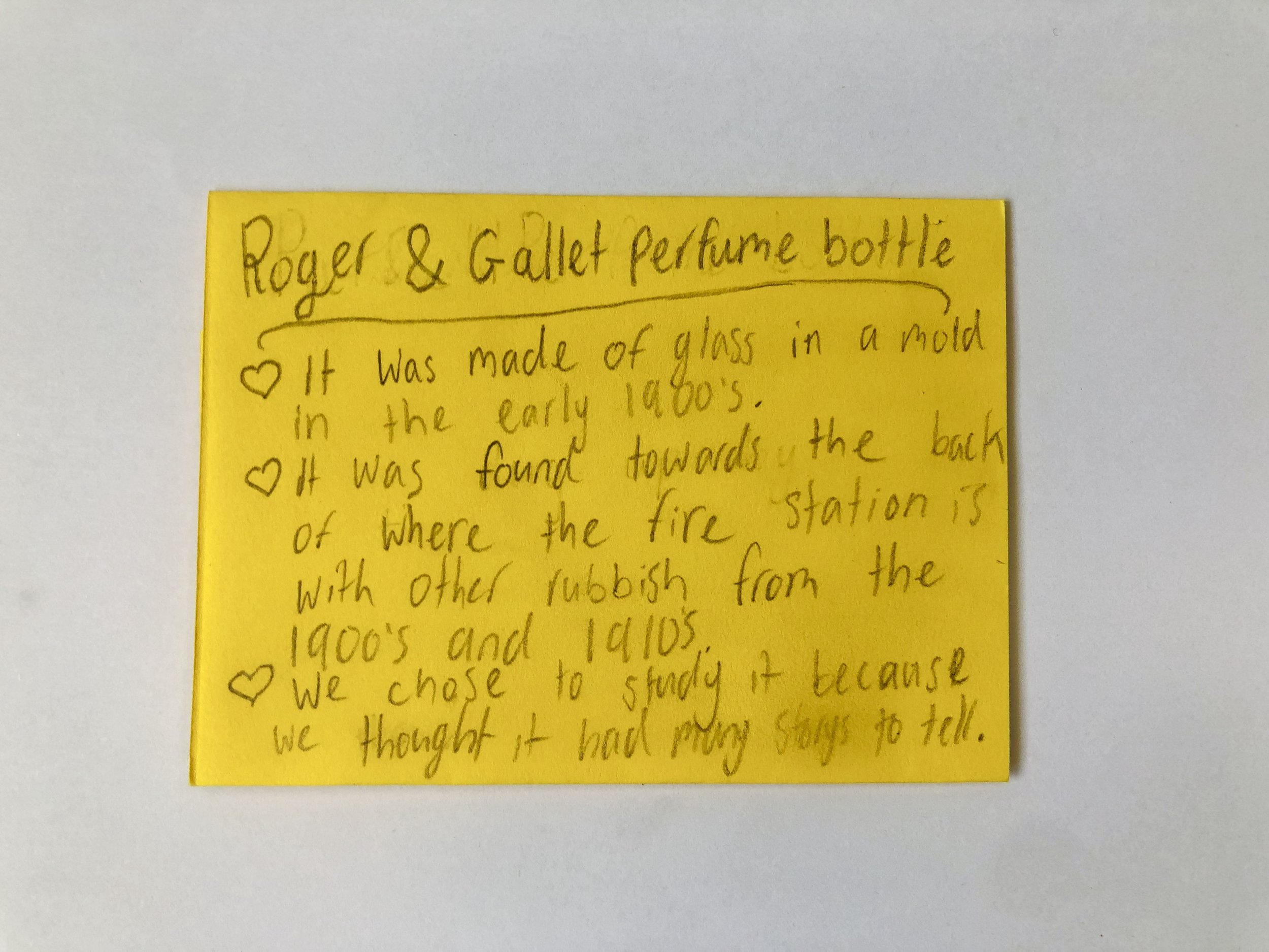 Yellow sticky note with handwritten text describing a perfume bottle made of glass from the early 1900s, found near a fire station, with some handwriting embellishments like hearts.