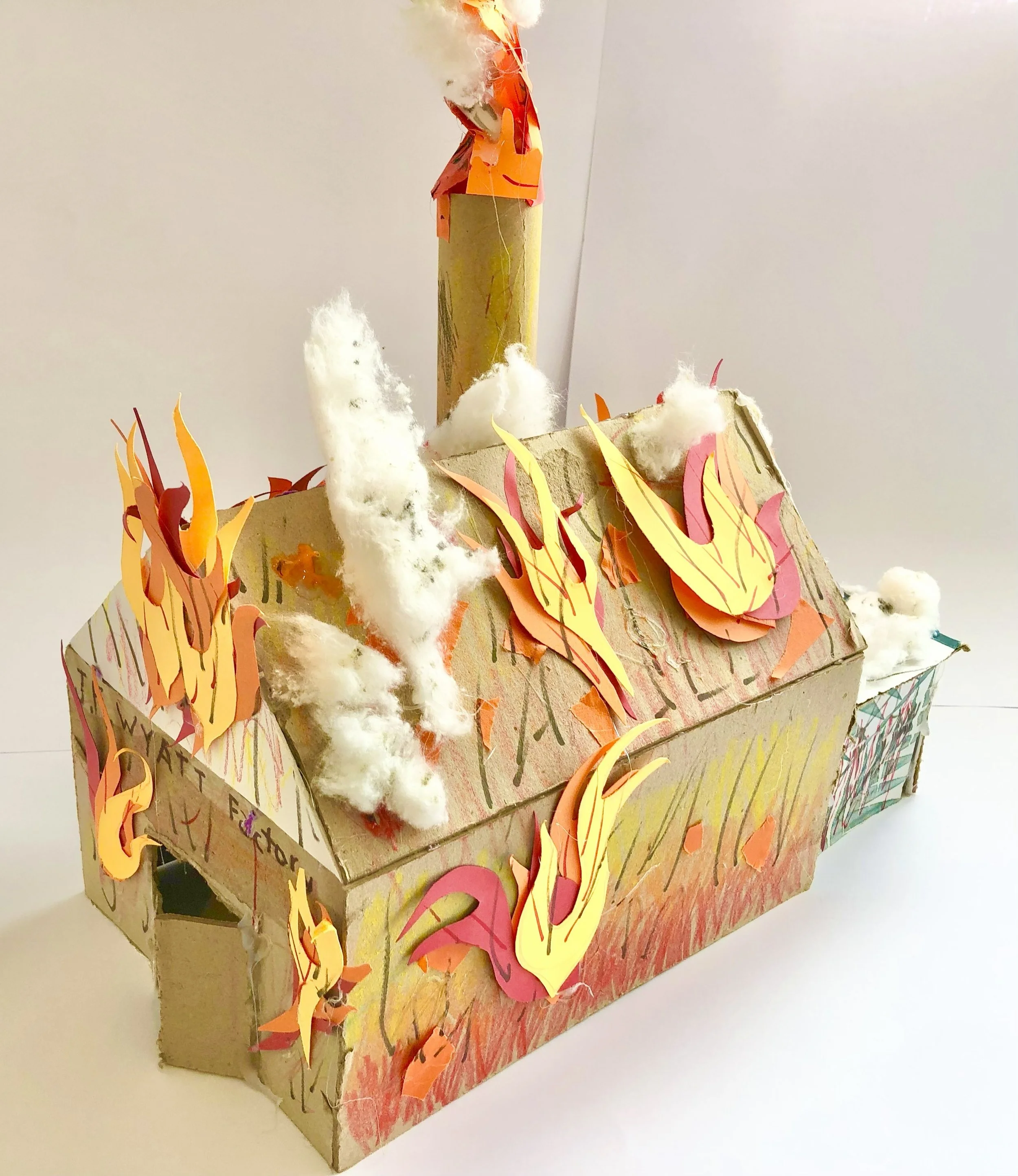 A paper craft model of a building on fire, made with cardboard, paper flames, cotton for smoke, and a paper chimney.