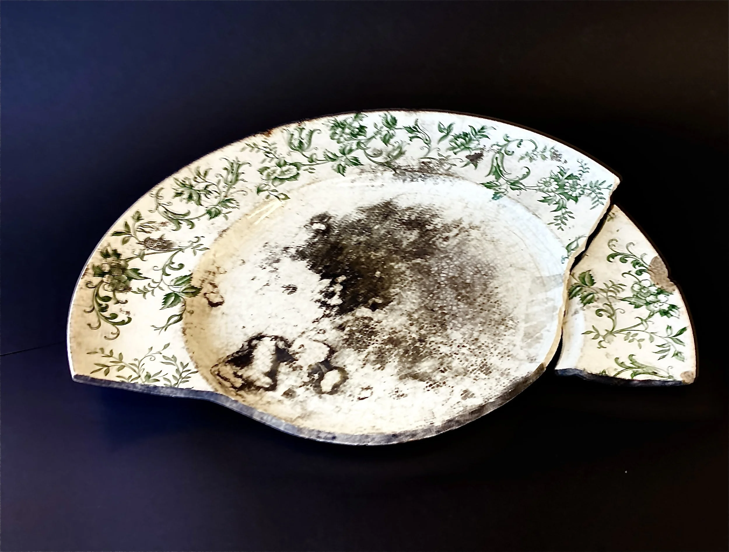 A broken ceramic plate with green floral patterns around the edges, dark stains and dirt on the inside surface, and a large chip missing from the rim, placed against a dark background.