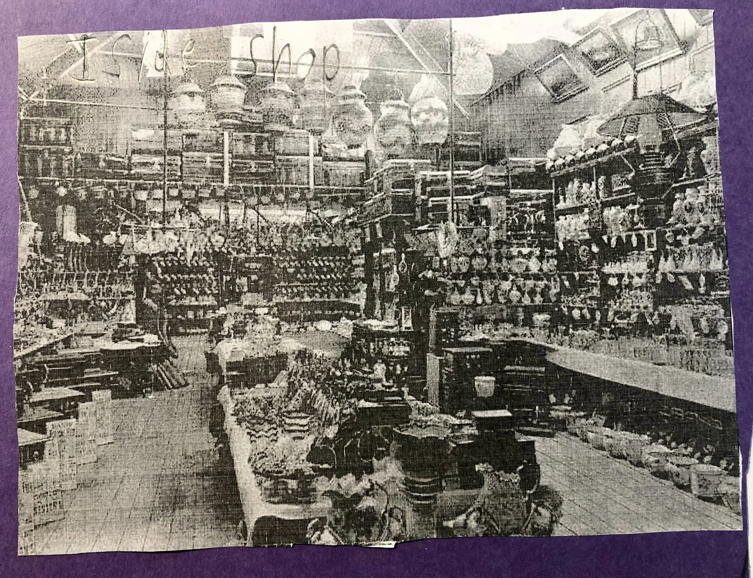 A black and white photo of a store aisle filled with various household items, including jars, ceramics, toys, and decorations, with shelves and hanging displays in the background.