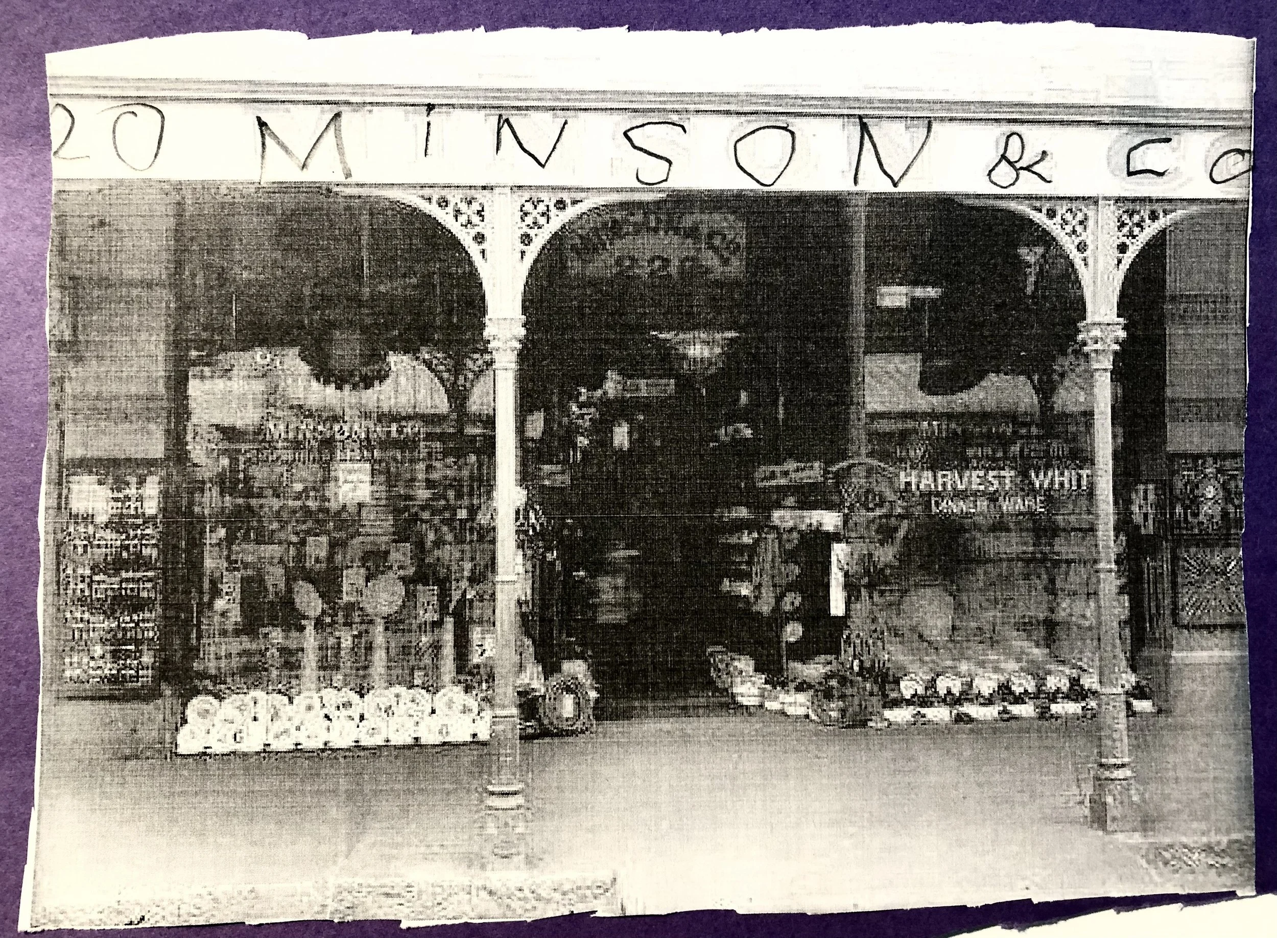 Black and white photo of a store entrance with the sign '20 MIN VISION & CO' at the top. The store sells various items including flowers, fruits, and vegetables, displayed outside. There are two decorative arches above the entrance and a sign for 'HARVEST WHITE FRUIT & VEGETABLES' visible inside.