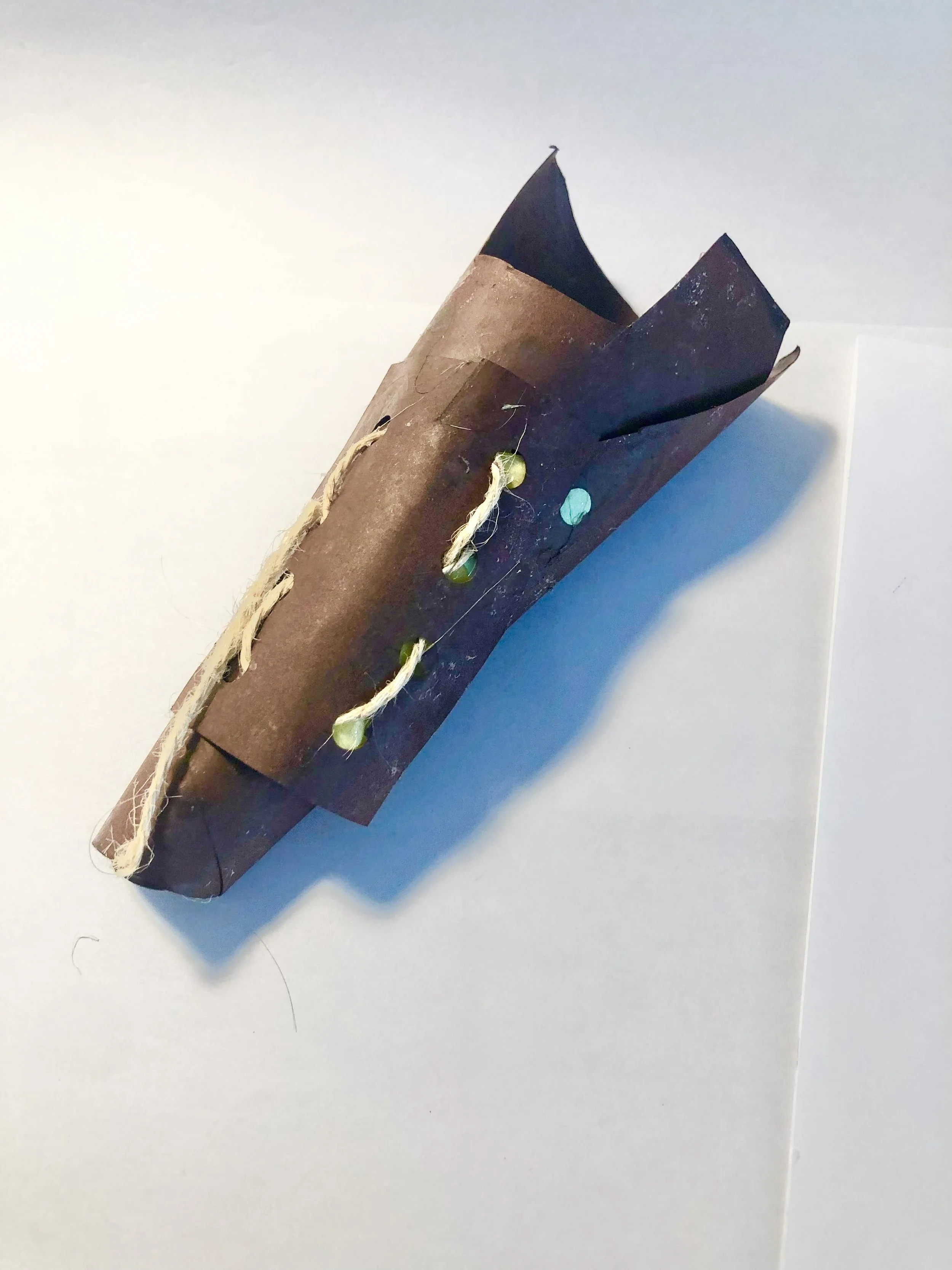 A homemade paper boat with dark brown and black colored paper, decorated with a string of small green elements, placed on a white surface with shadows.