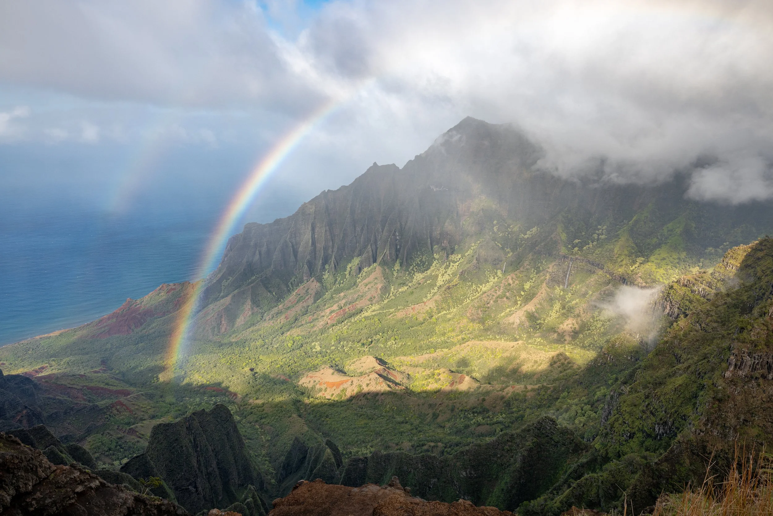 A scenic landscape featuring a lush green valley with mountains partially covered by clouds, and a rainbow arching through the sky over the mountains and ocean.