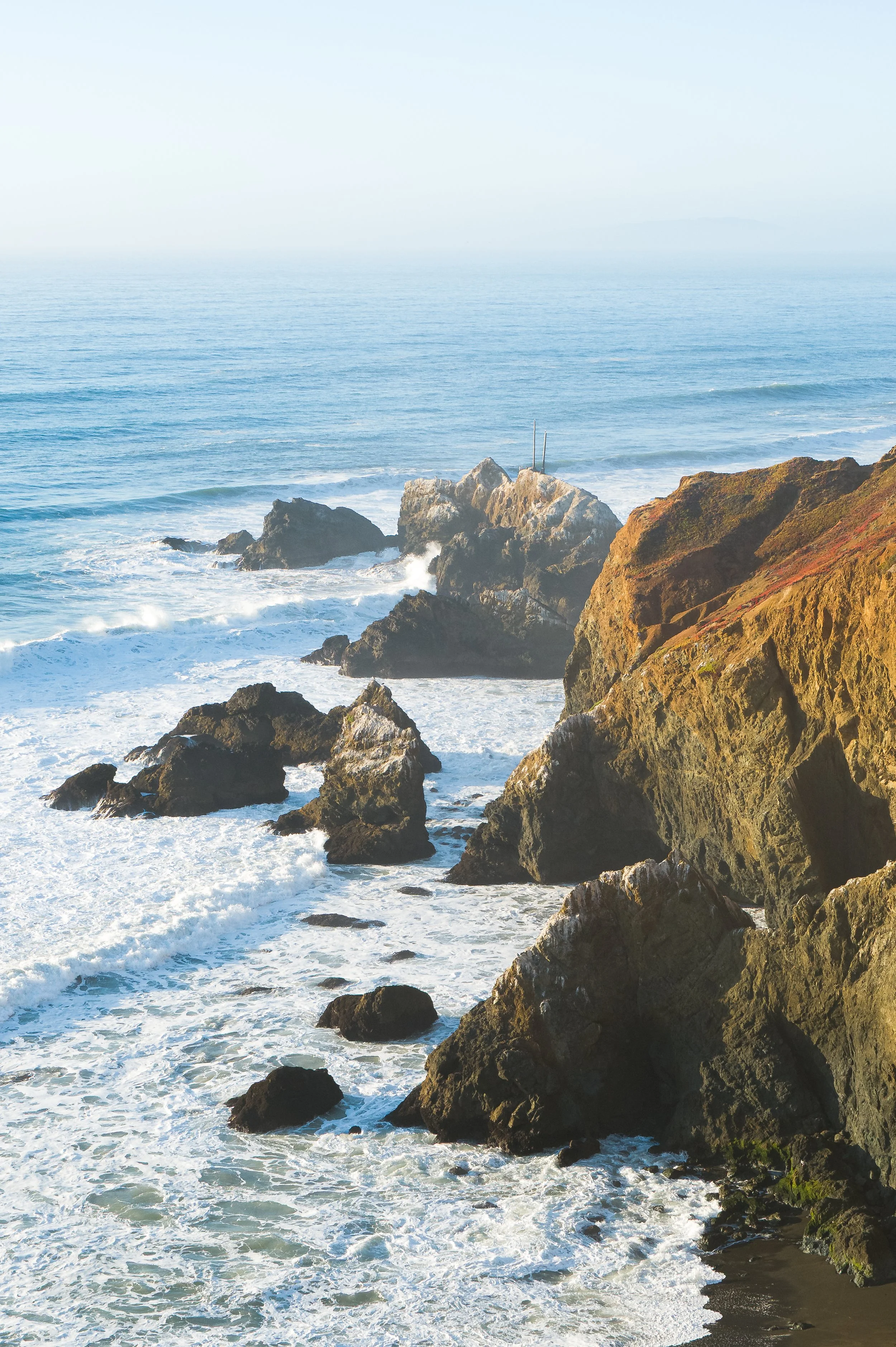 Ocean waves crashing against large rocks along a coastal cliff at golden hour along the California coast.