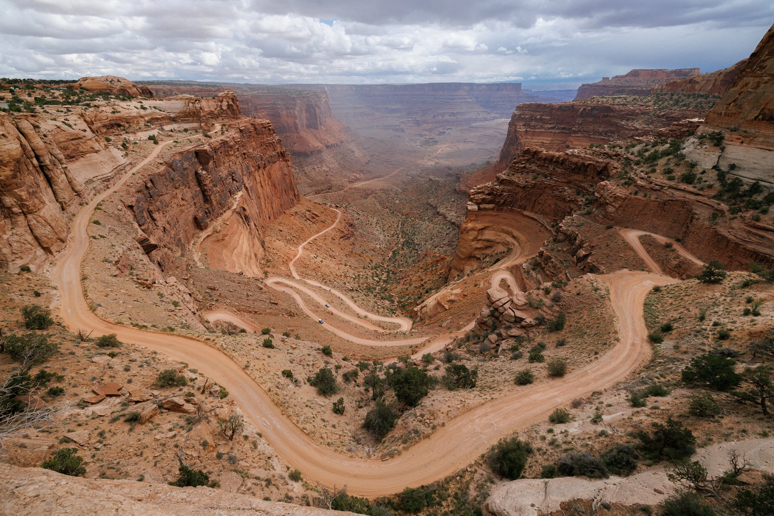 A panoramic view of the Grand Canyon with winding dirt roads, layered red and brown rock formations, sparse desert vegetation, and a cloudy sky overhead.