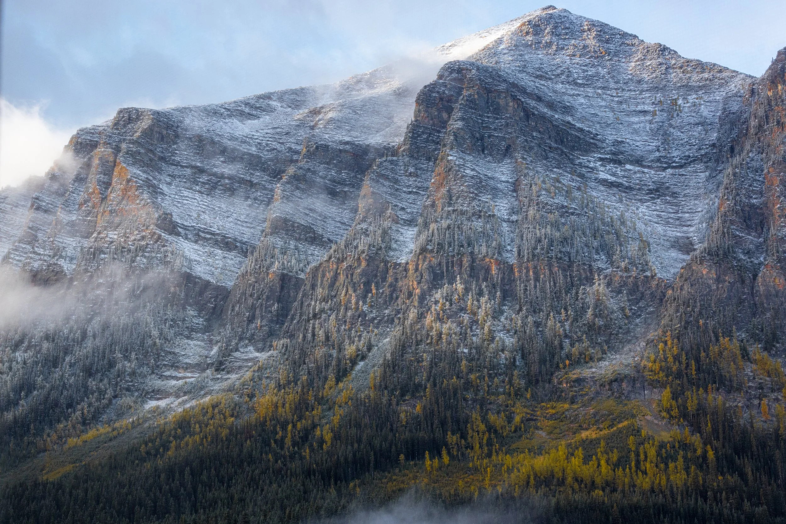 Snow-capped mountain with rocky cliffs and evergreen trees at the base, some mist or fog surrounding the area in Banff National Park.
