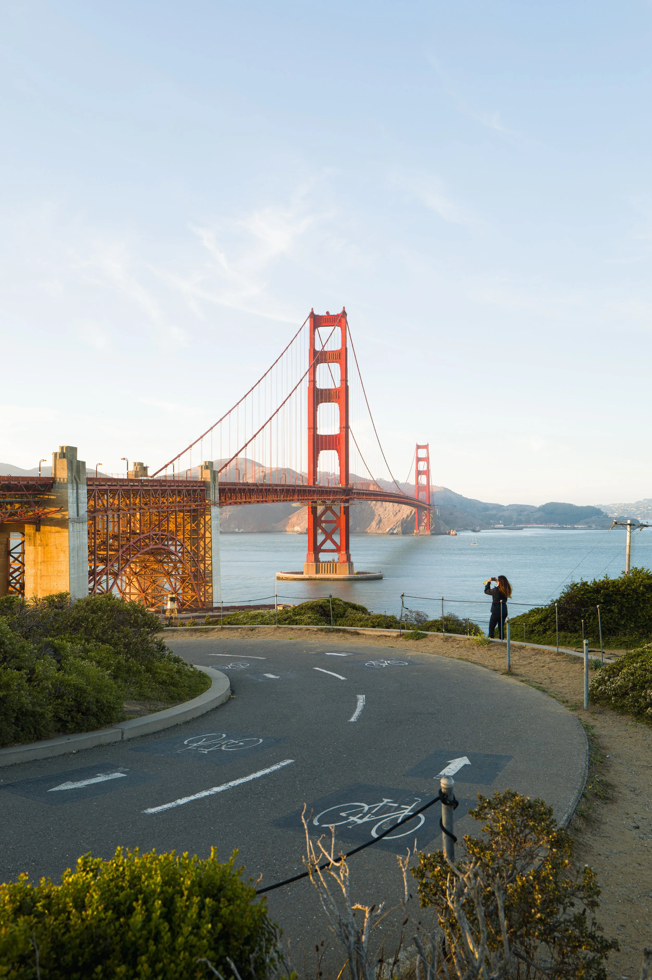 View of the Golden Gate Bridge in San Francisco, with a person taking a photo on the viewpoint, and cycling lane markings in the foreground.