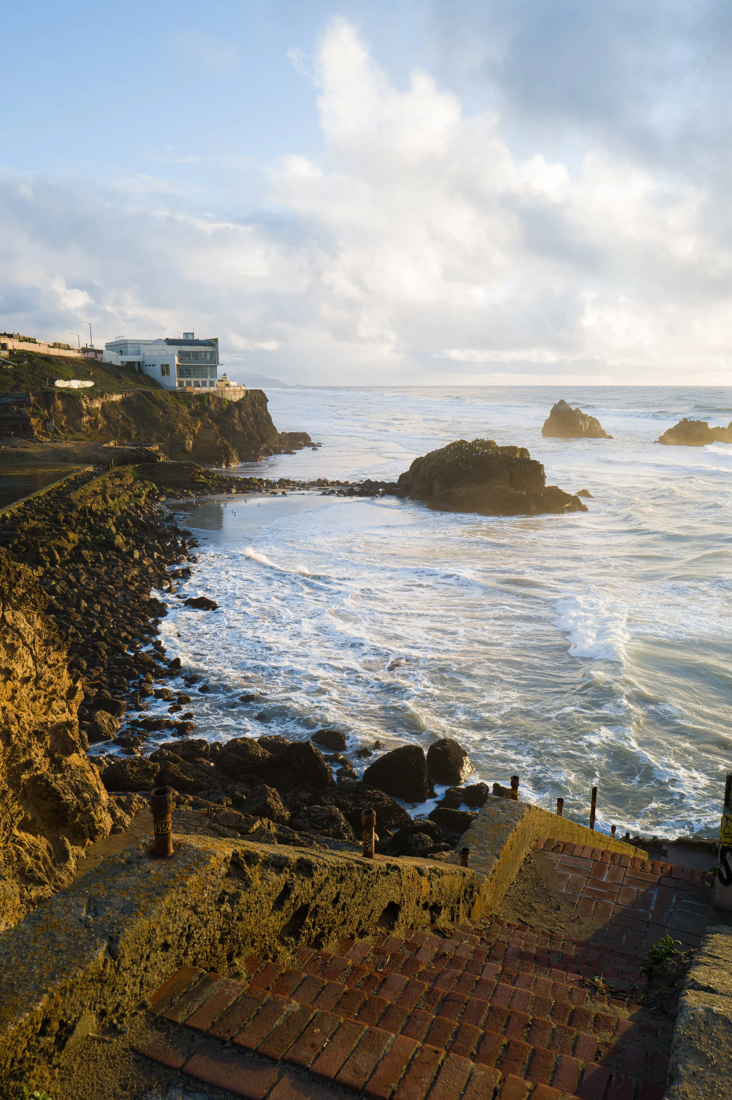 View of a rocky coastline with waves crashing against the rocks, a building on the cliff, and a partly cloudy sky at the Sutro Baths in San Francisco.