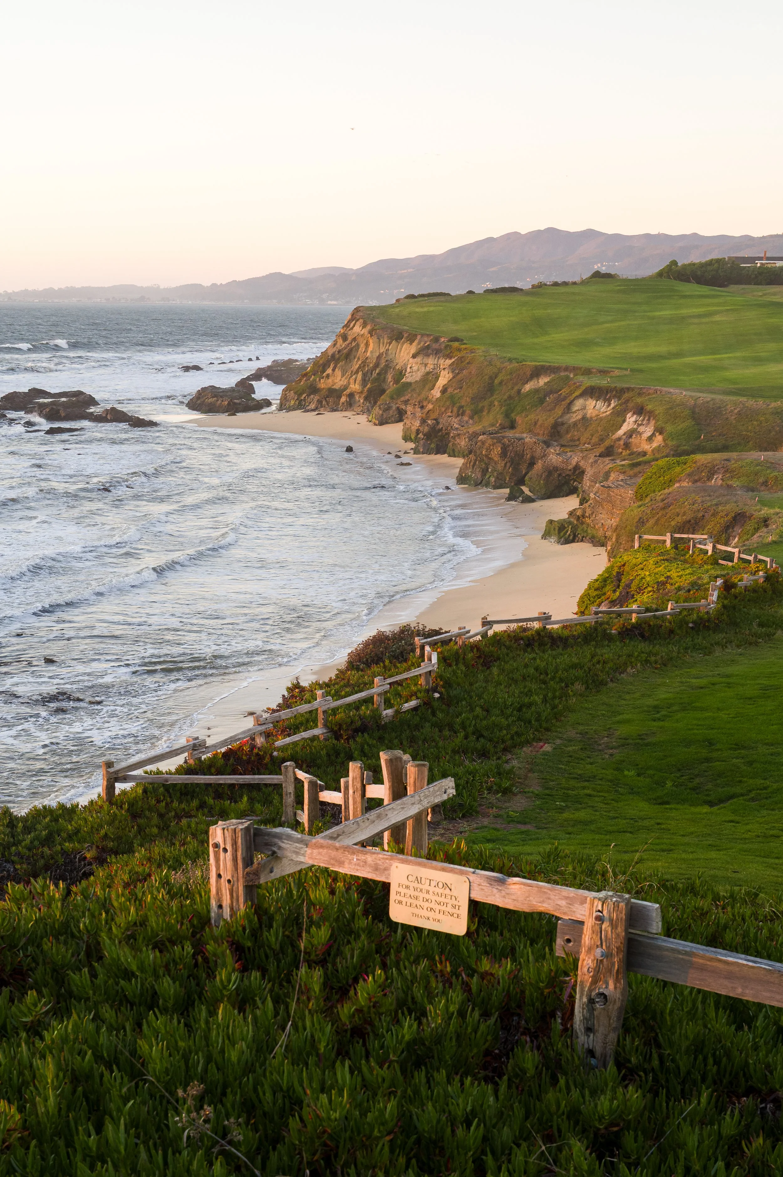 Scenic view of the coastline with sandy beach, rocky cliffs, and green grassy areas, with wooden fences and a caution sign near the edge in Half Moon Bay, CA.