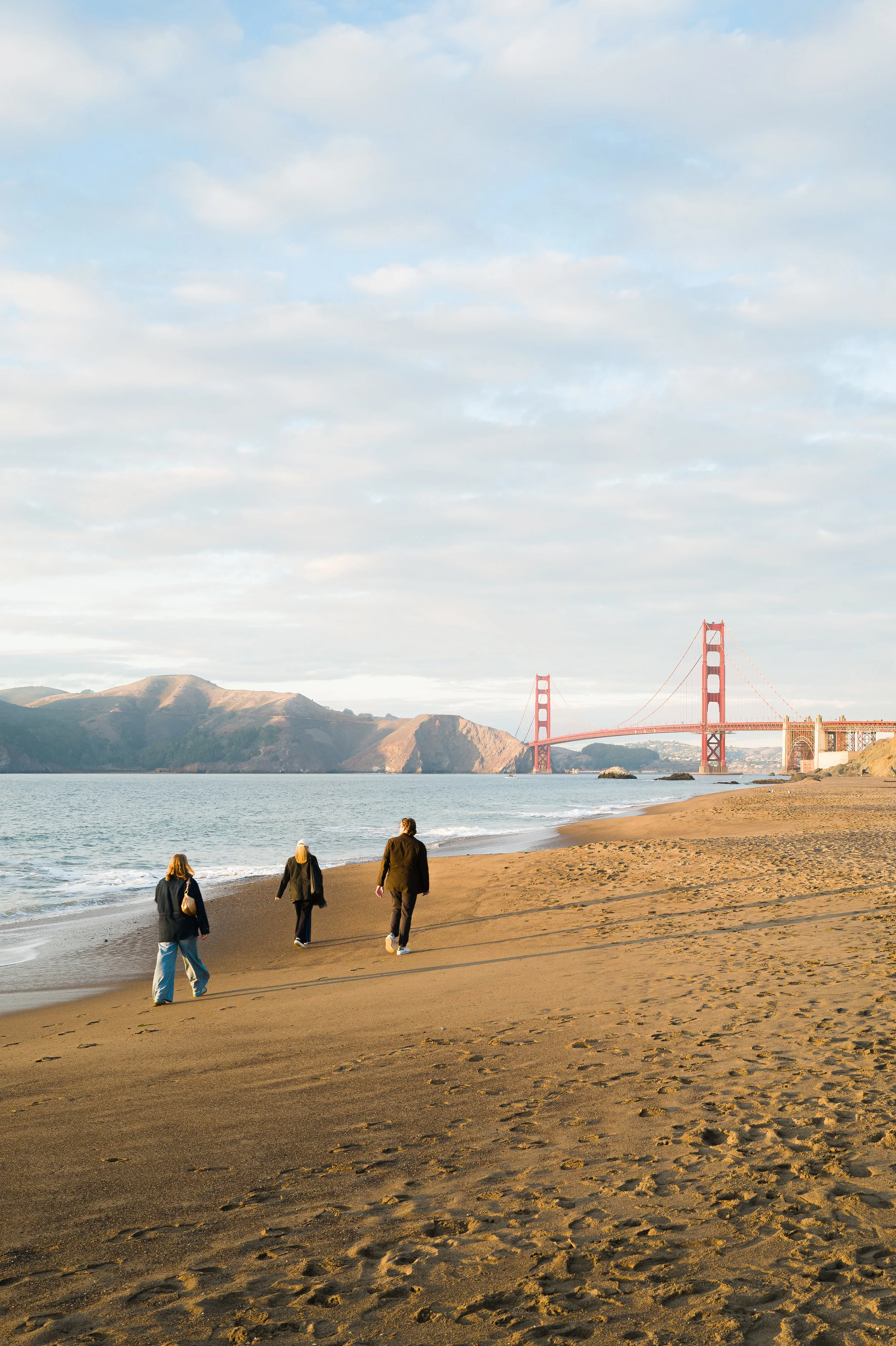 People walking on a sandy beach near the Golden Gate Bridge with hills in the background under a partly cloudy sky.
