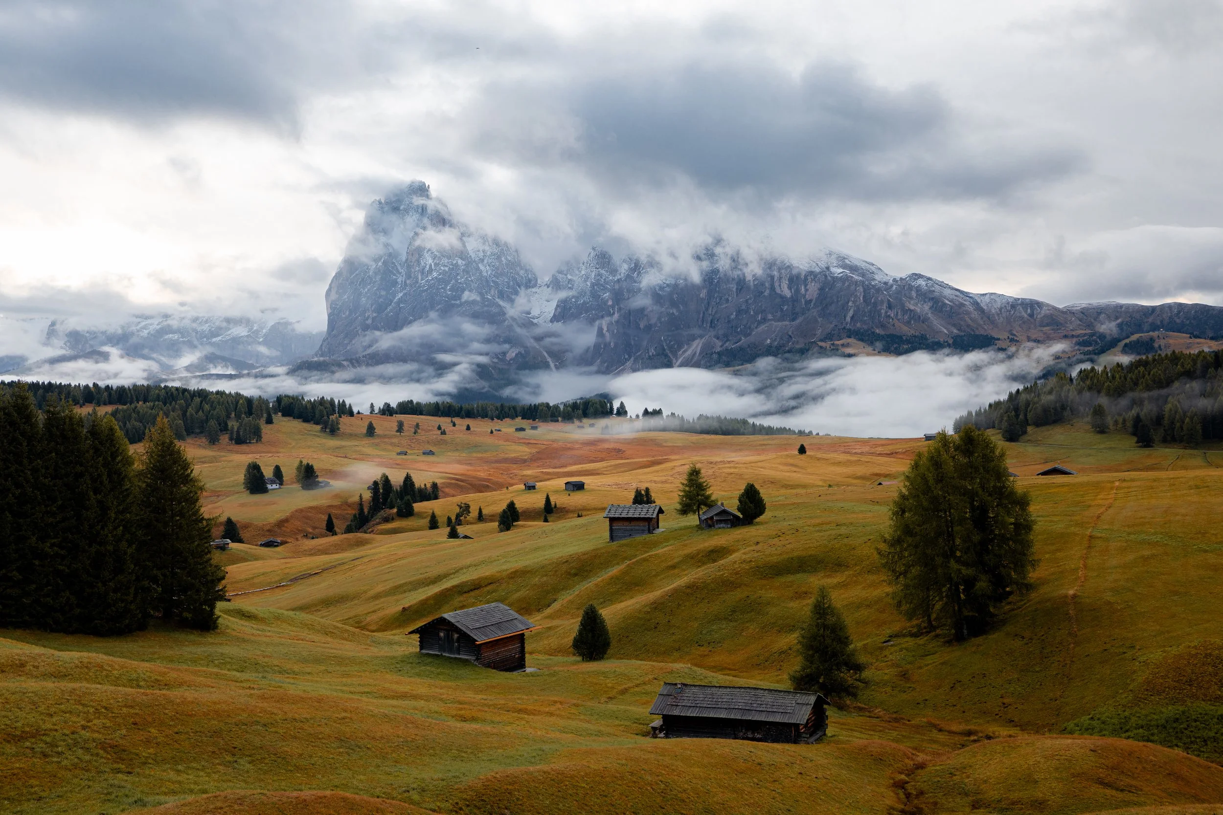 A scenic landscape of rolling green hills with small wooden cabins, scattered trees, and misty mountains in the background under a cloudy sky in the Italian Dolomites.