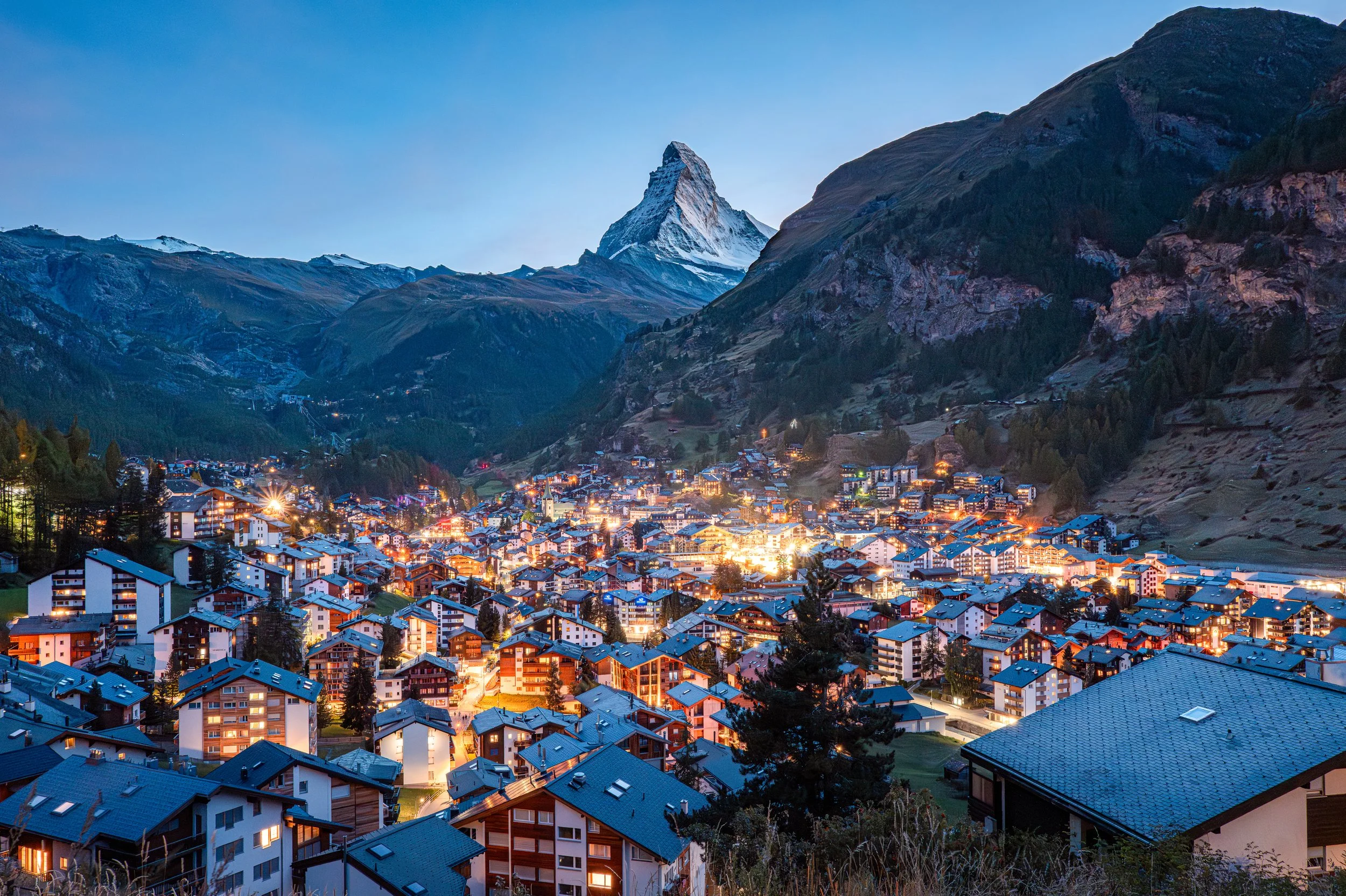 Nighttime view of a mountain village with illuminated buildings, set against the backdrop of snow-capped mountains and the Matterhorn in the distance.