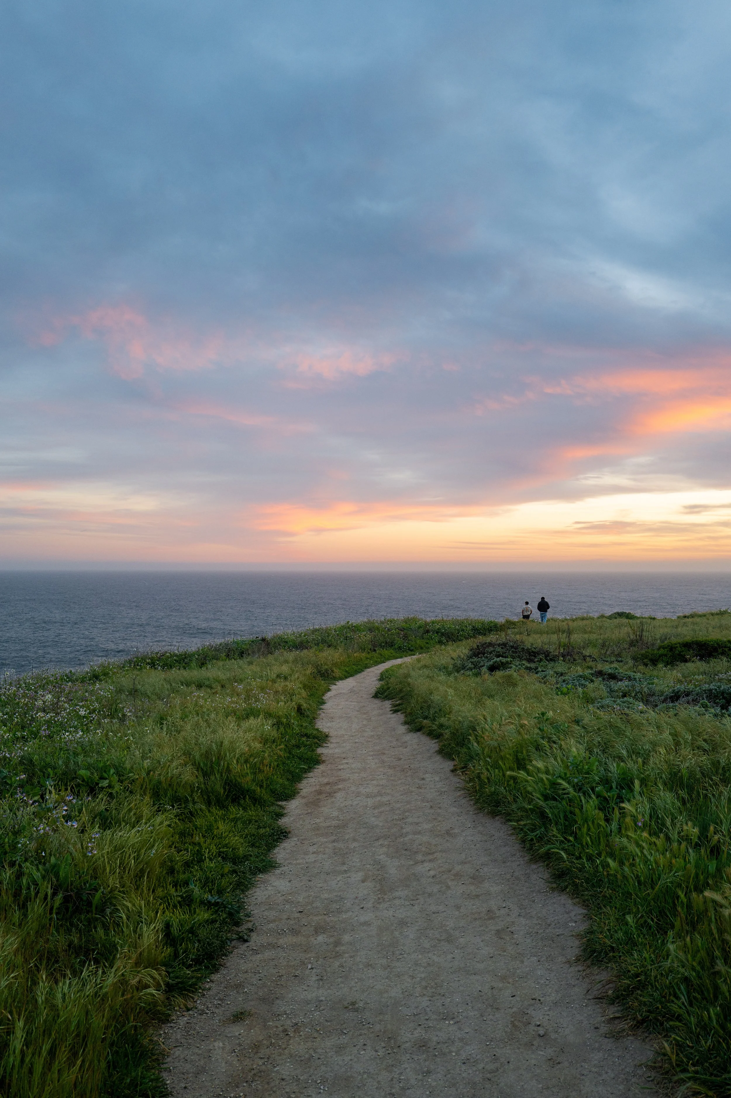A dirt path through green grass and bushes leading to two people standing at the edge of a cliff overlooking the ocean during sunset, with a sky filled with clouds and pinkish hues.