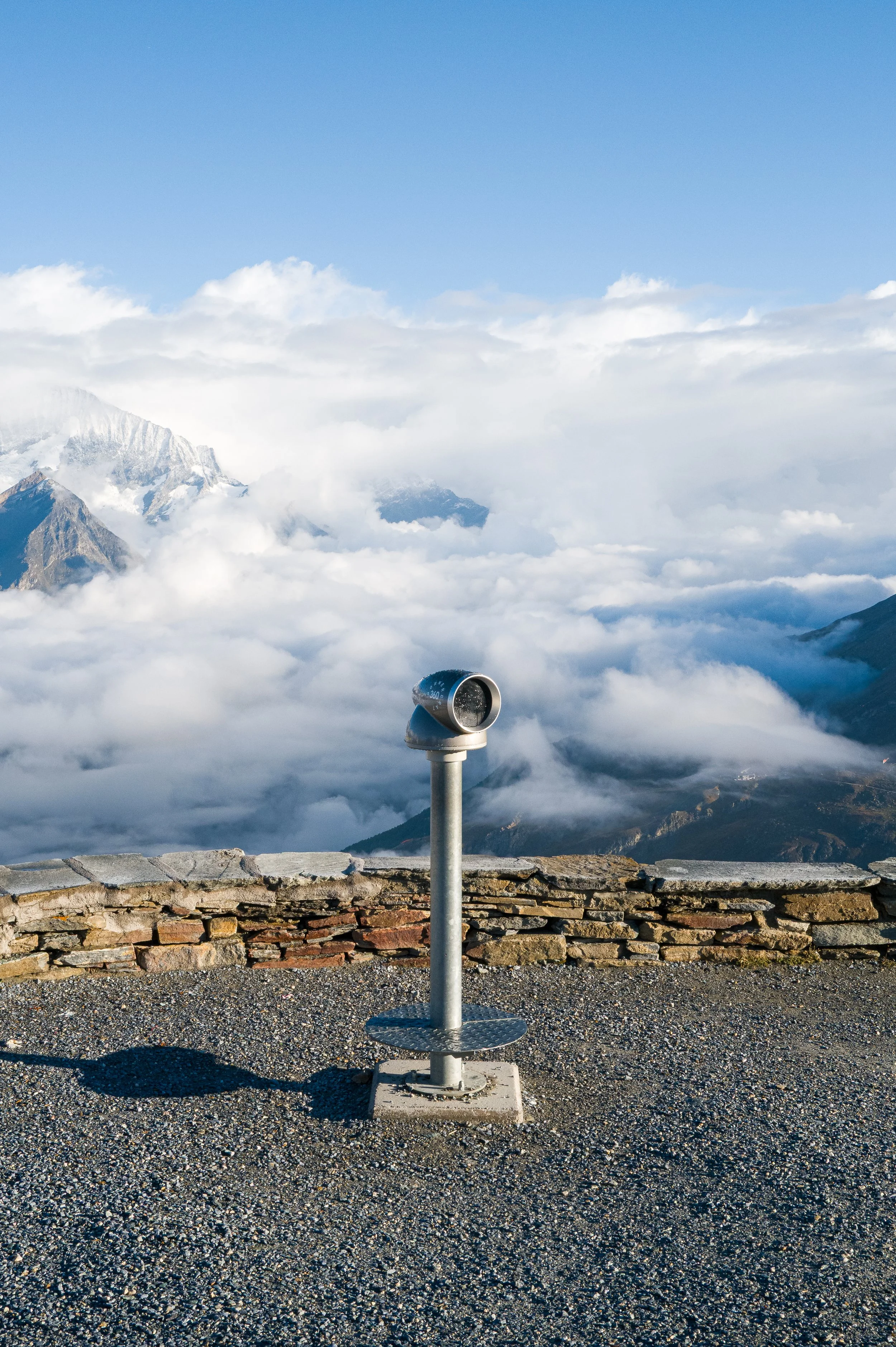 Tourist viewing telescope on a mountain overlook with clouds, snow-capped peaks, and rugged terrain in the background.