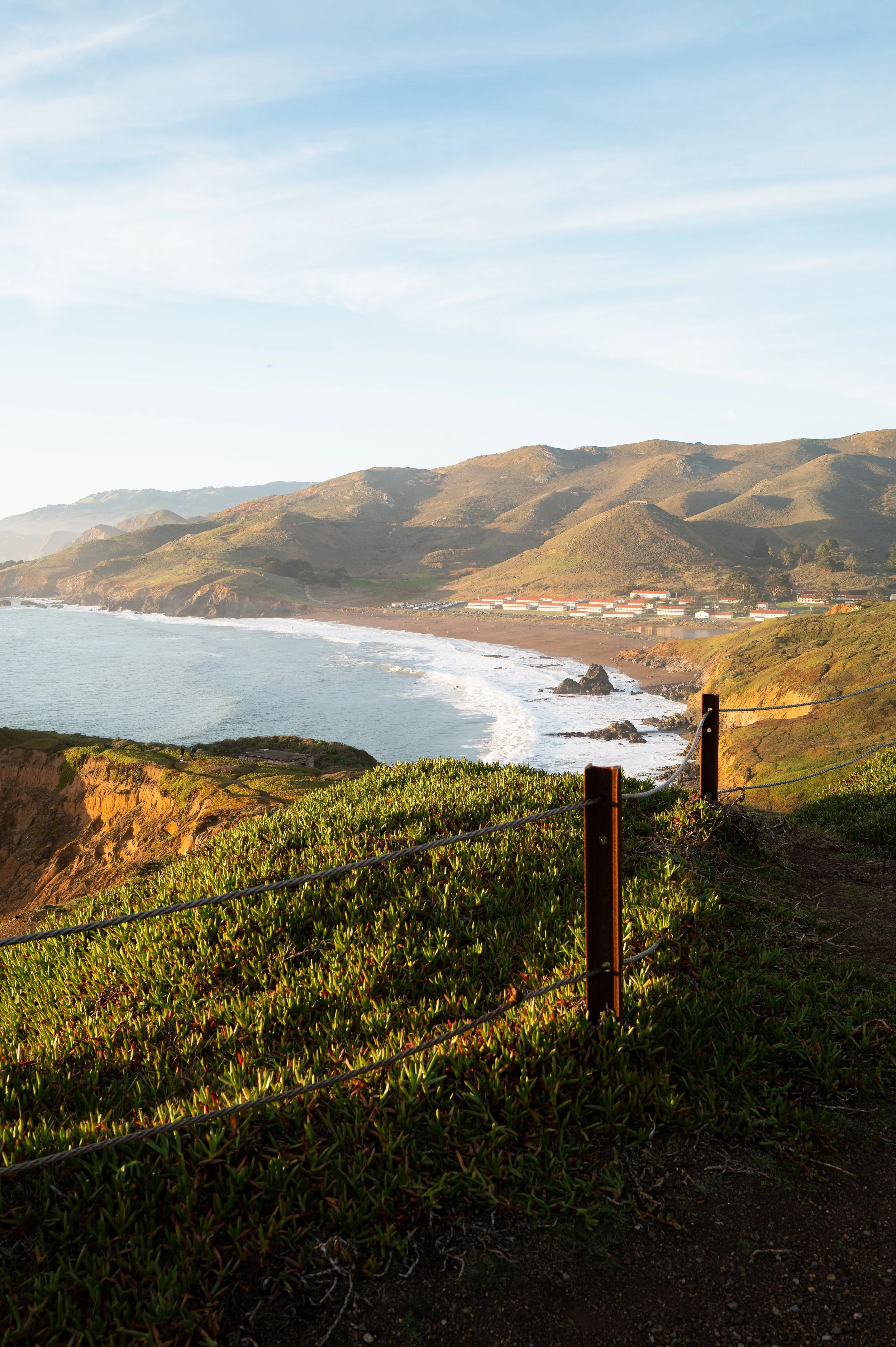 Coastal landscape with cliffs, vegetation, and a view of the ocean and distant mountains, under a clear sky.