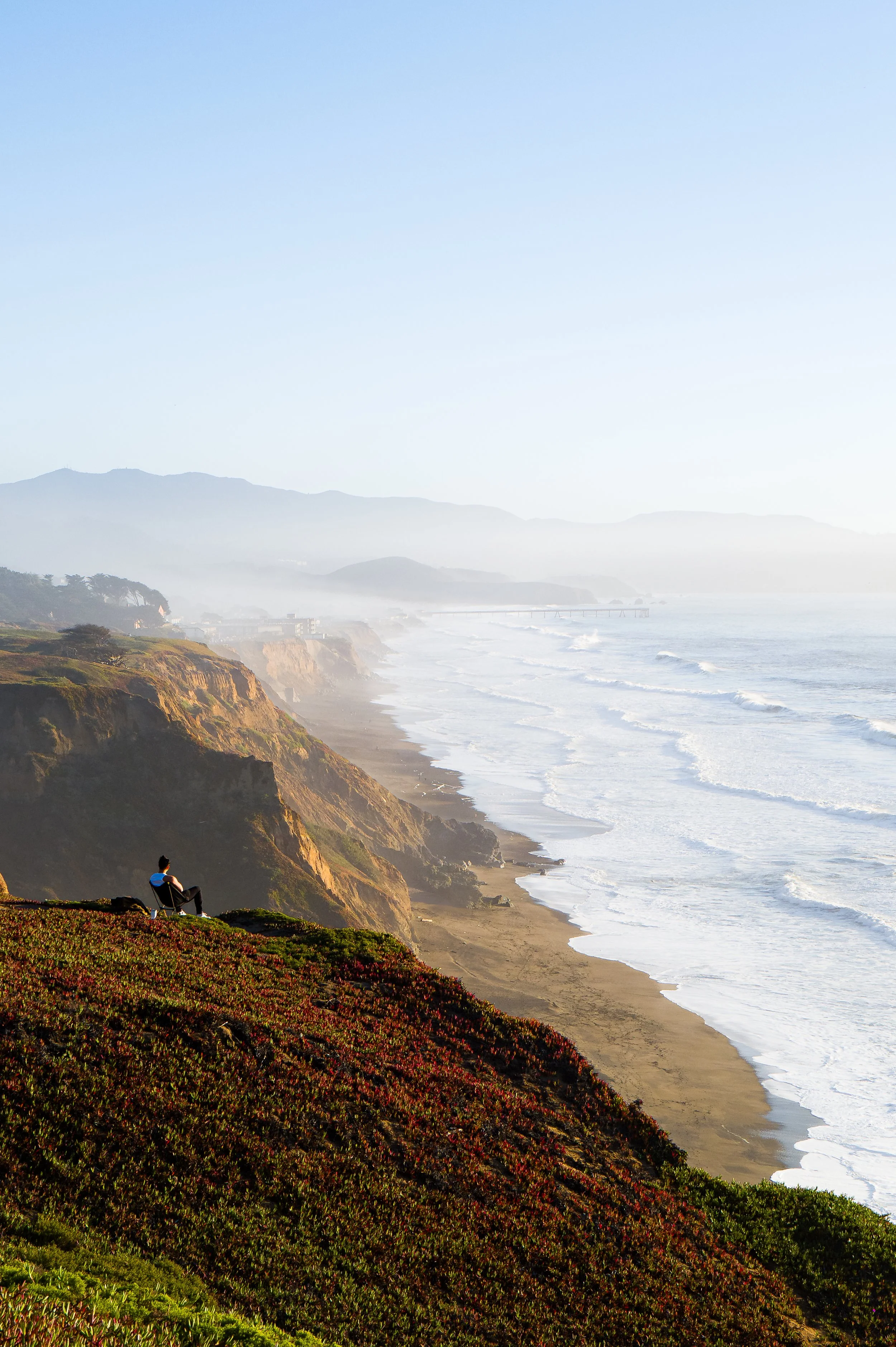 A person sitting on a bench on a grassy hill overlooking the coastline with cliffs, a sandy beach, and the ocean extending to the horizon, with distant mountains and a pier in the background on a clear day.