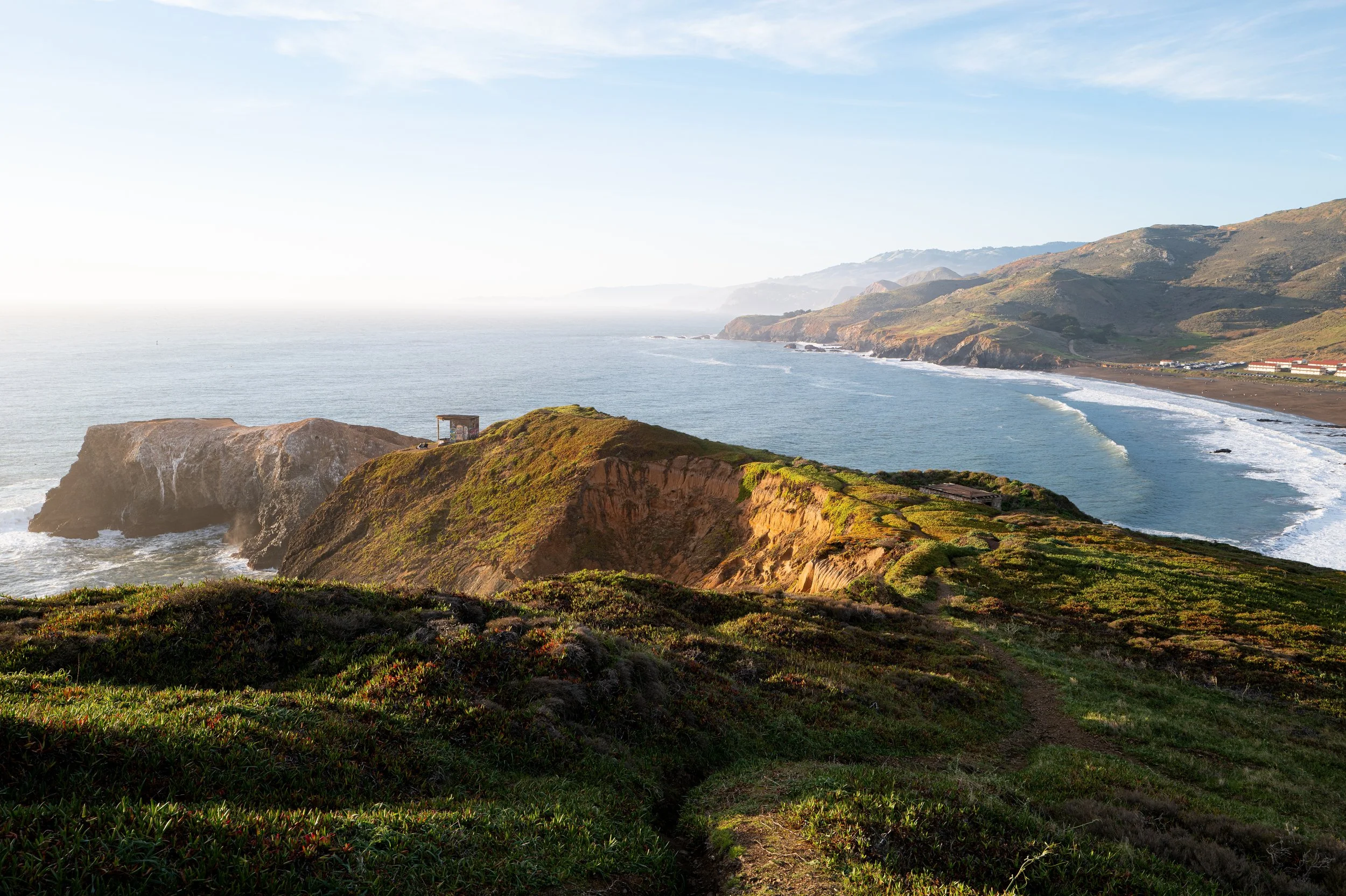 A coastal landscape with green hills leading to rocky cliffs and a sandy beach, with the ocean to the left and mountains in the background under a blue sky along the California Coast.