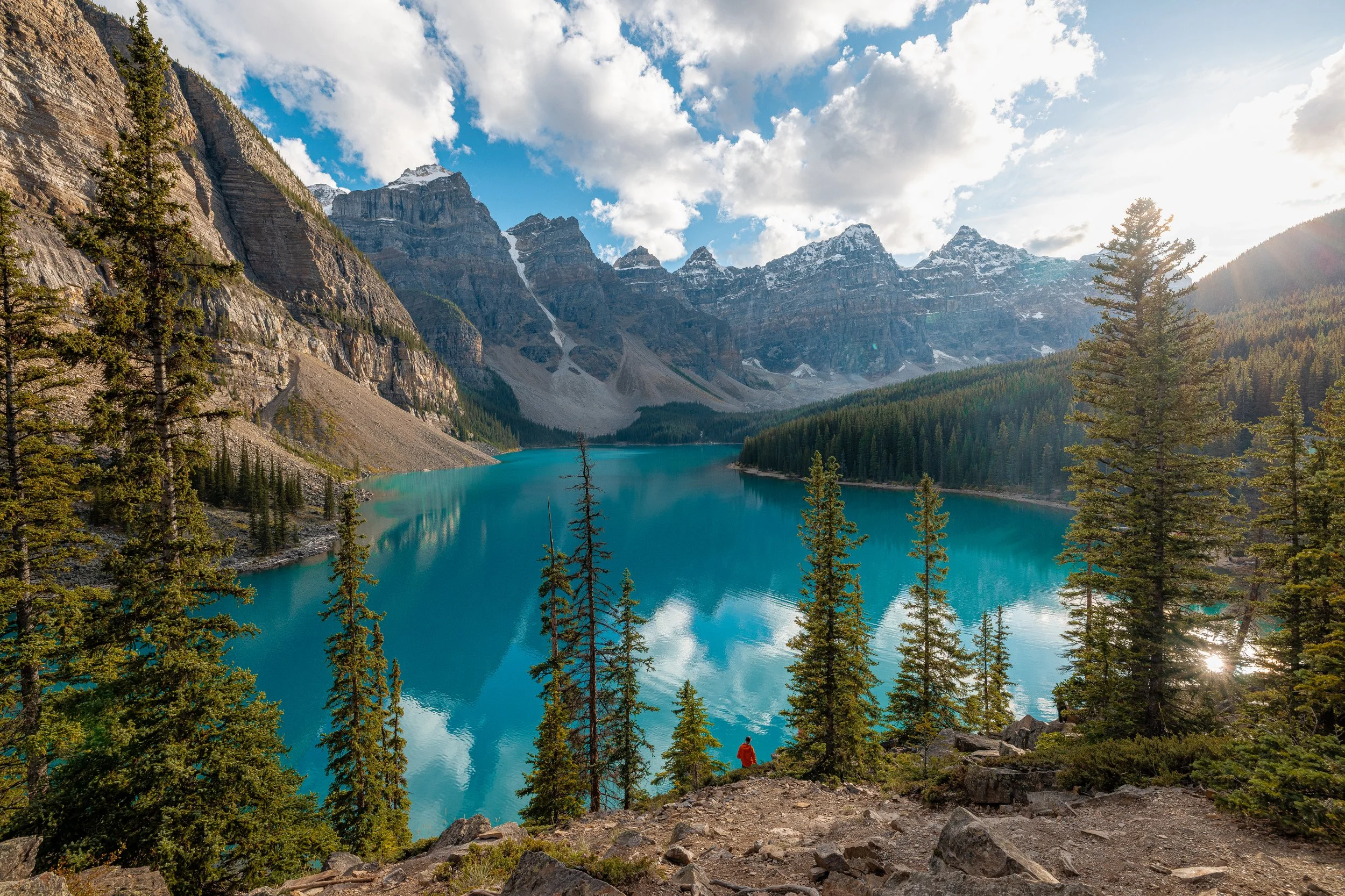 Scenic view of a turquoise mountain lake surrounded by pine trees, rocky terrain, and high snow-capped mountains under a partly cloudy sky, with sunlight reflecting off the water at Lake Louise in Banff National Park.