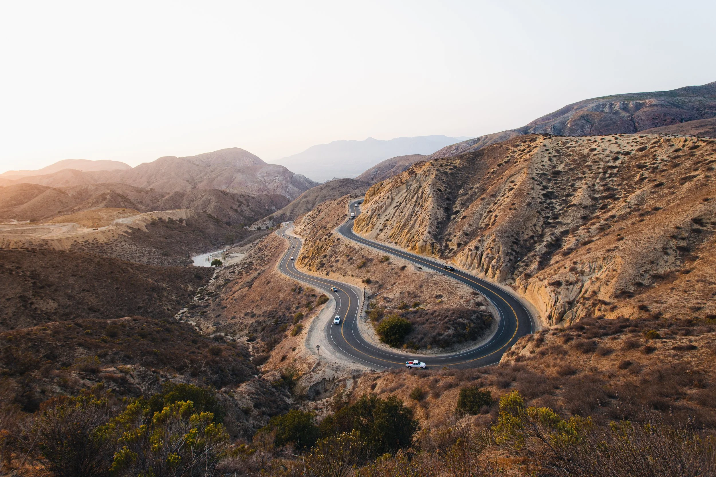 Curving mountain road with several vehicles driving through a dry, hilly desert landscape at sunset in the Los Angeles mountains.