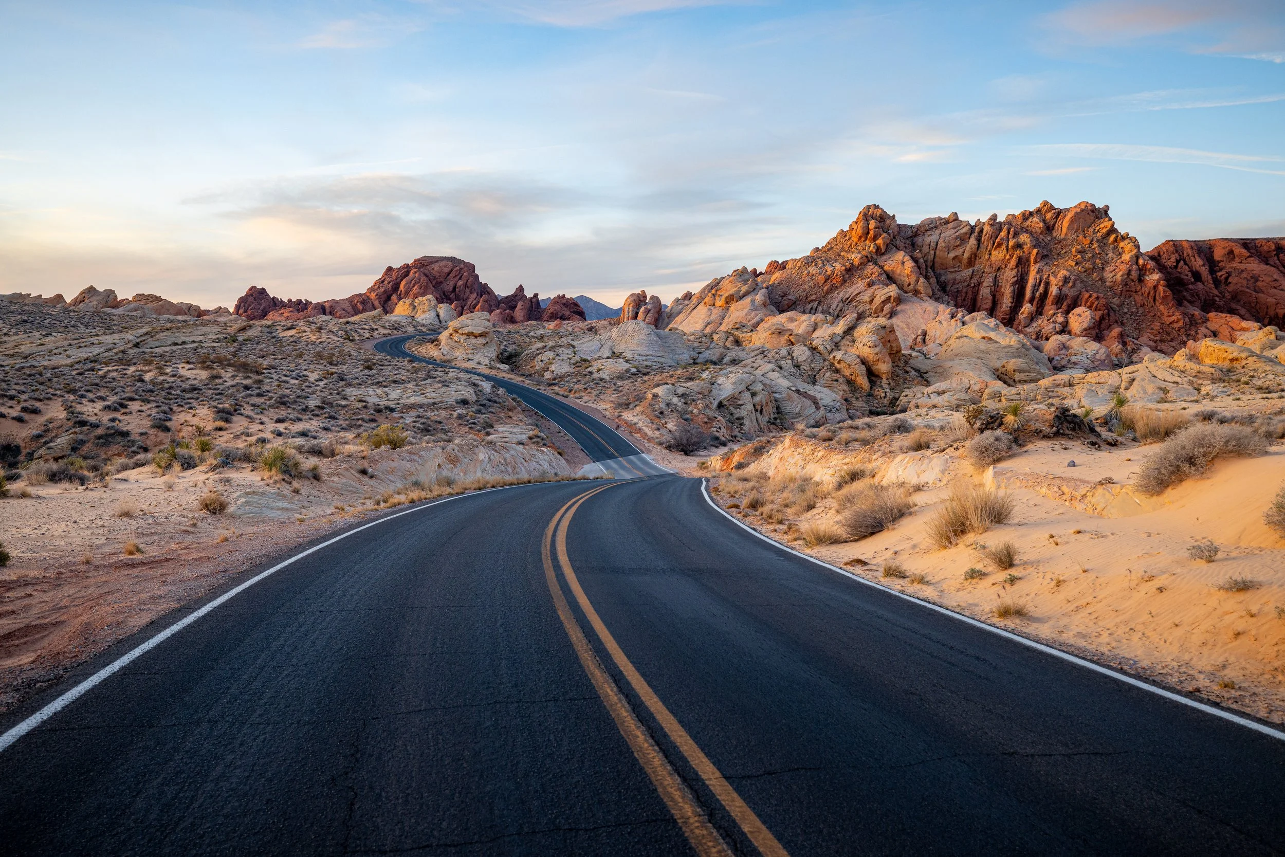 Winding asphalt road through desert landscape with rocky hills and sparse vegetation under a sky with soft clouds.