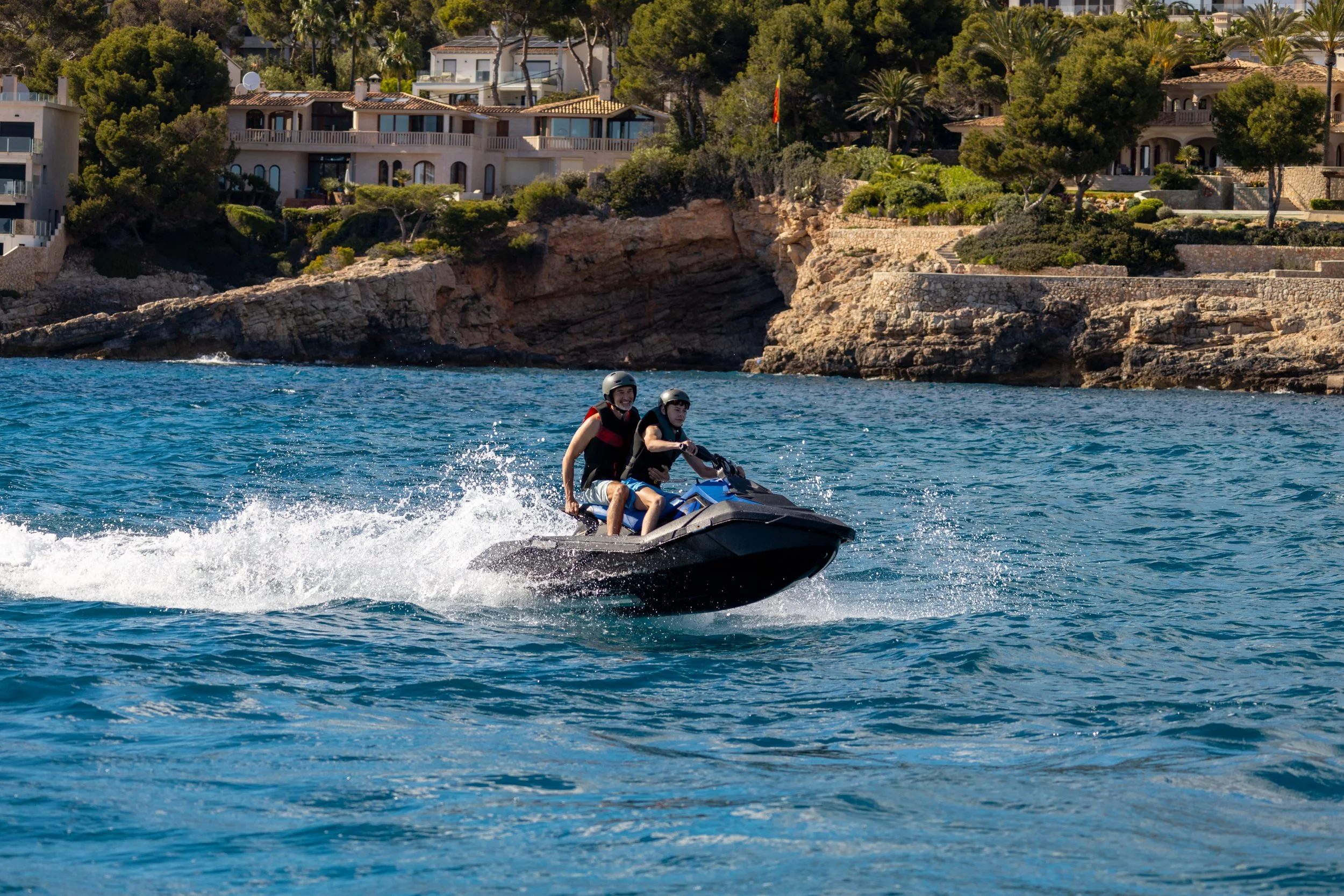 Two people wearing helmets riding a jet ski on a body of water near rocky shoreline with houses and trees in the background.