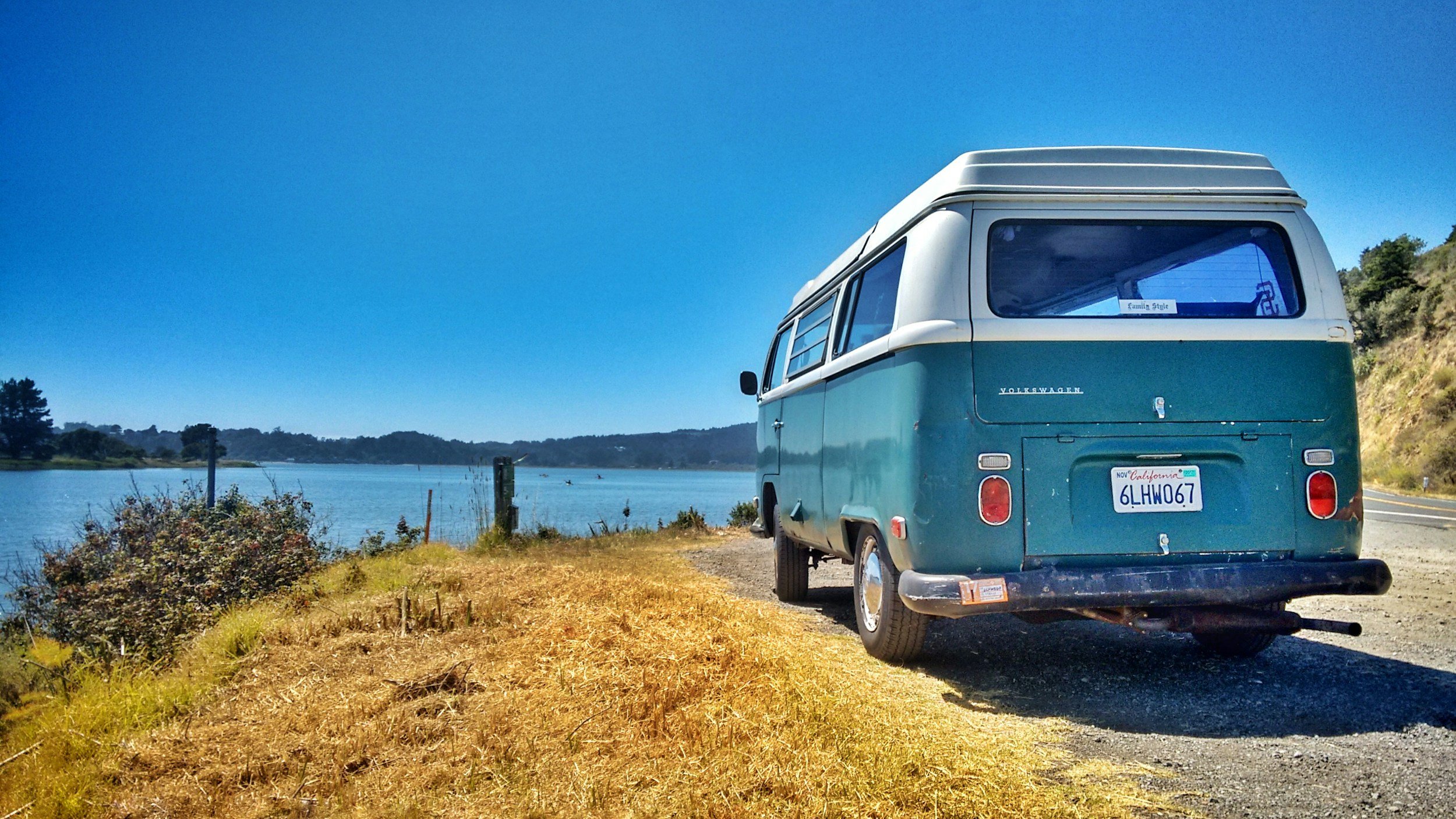 A teal and white vintage Volkswagen camper van parked beside a lake on a sunny day, with trees and hills in the background.