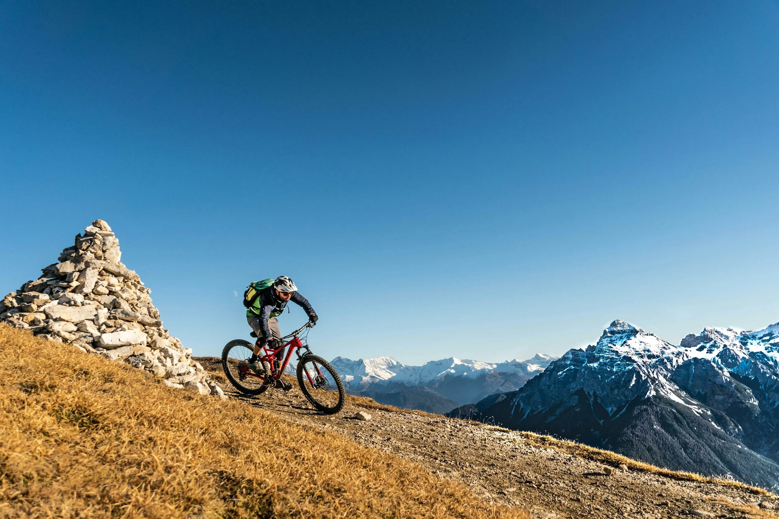 A mountain biker riding downhill on a trail with a view of snow-capped mountains in the background during daytime.