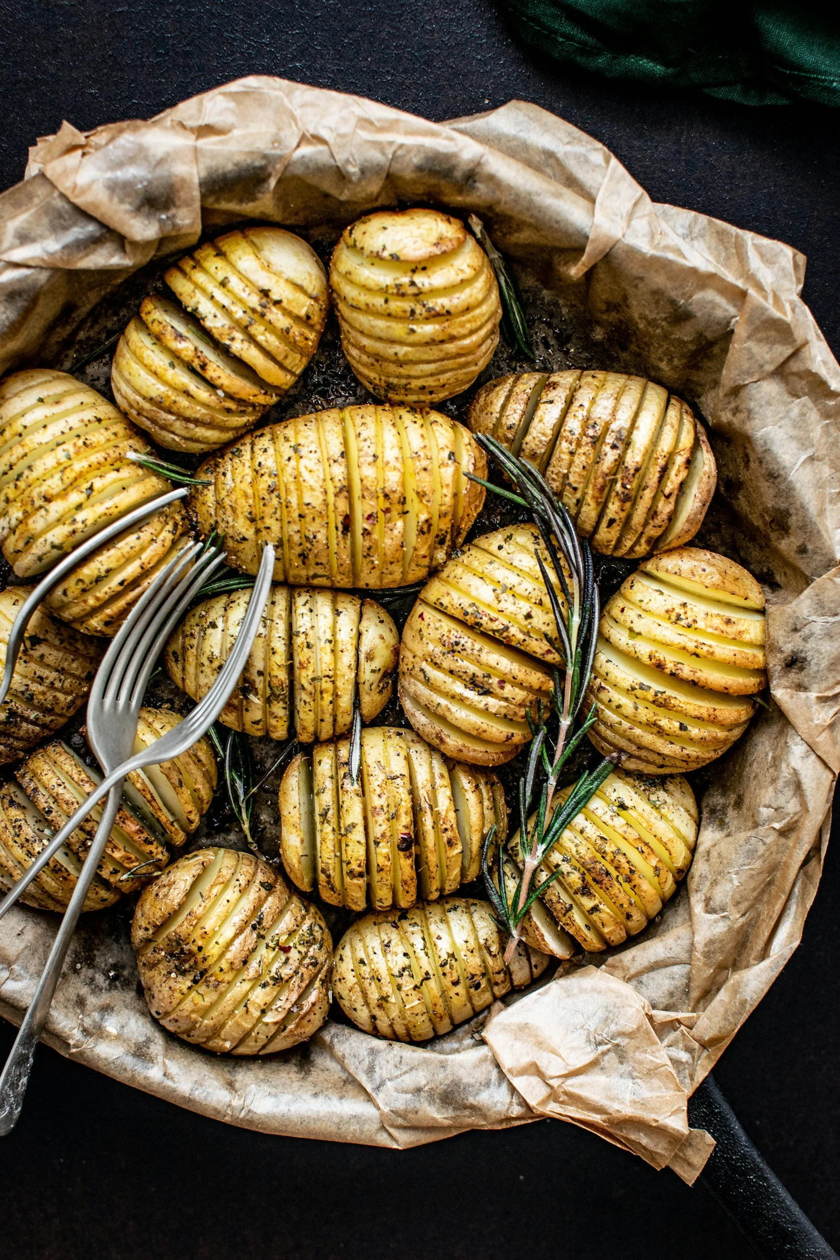 Hasselback potatoes seasoned with herbs and spices, garnished with rosemary sprigs, in a parchment-lined baking dish.