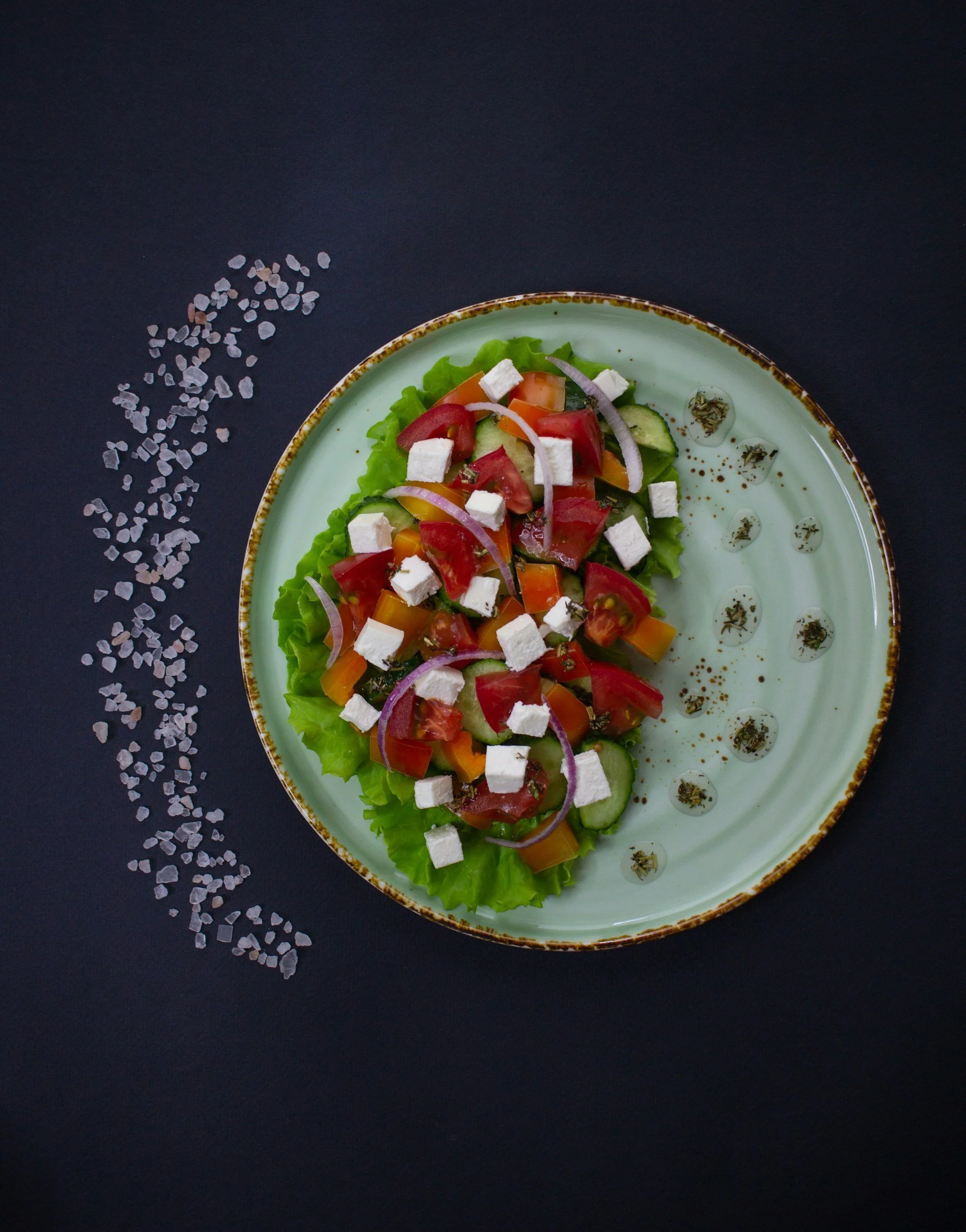 Fresh salad with lettuce, tomatoes, cucumbers, red onions, feta cheese, and herbs on a decorative light green plate against a dark background, with salt crystals scattered nearby.