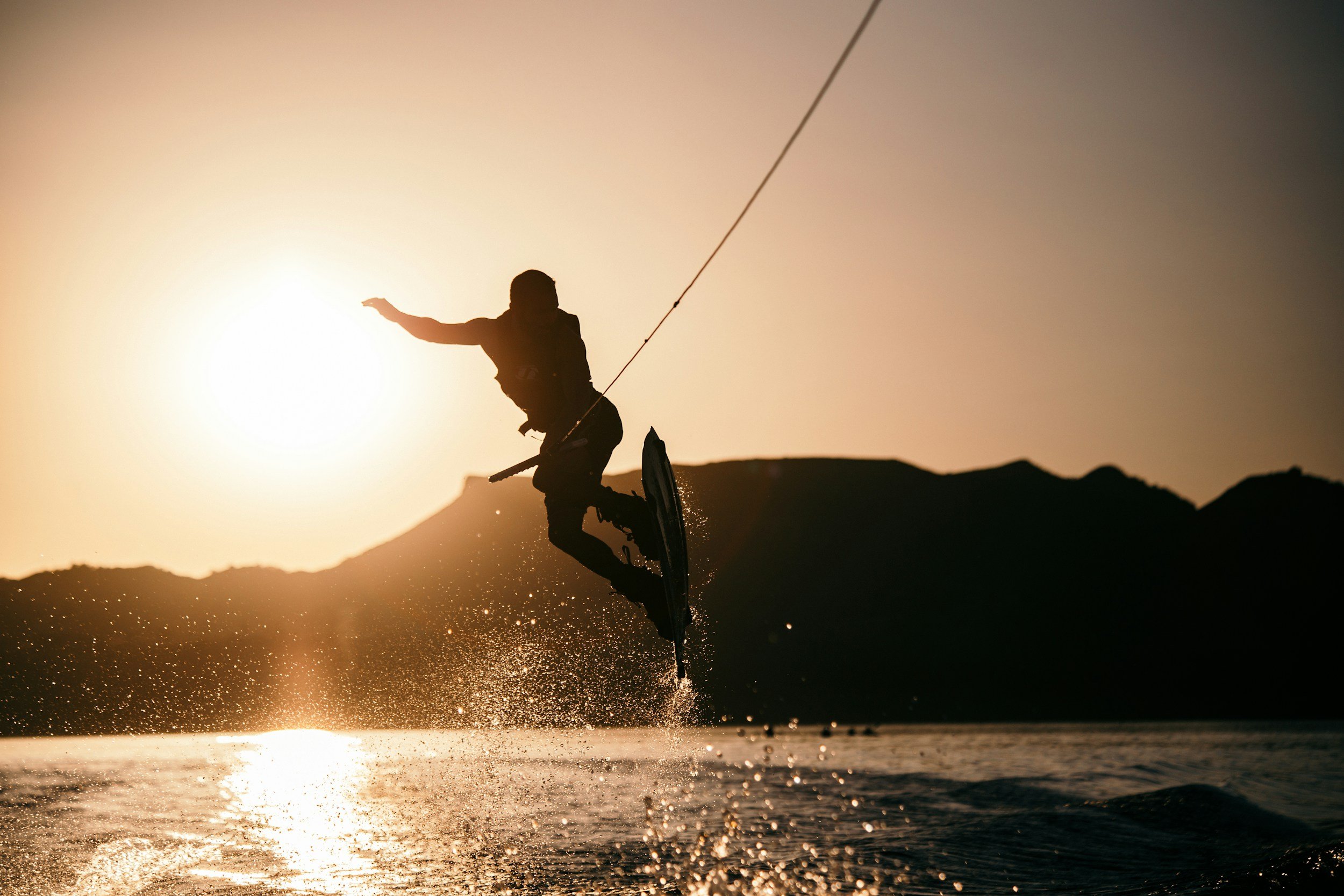 A person wakeboarding on the water at sunset, silhouetted against the sun with mountains in the background.