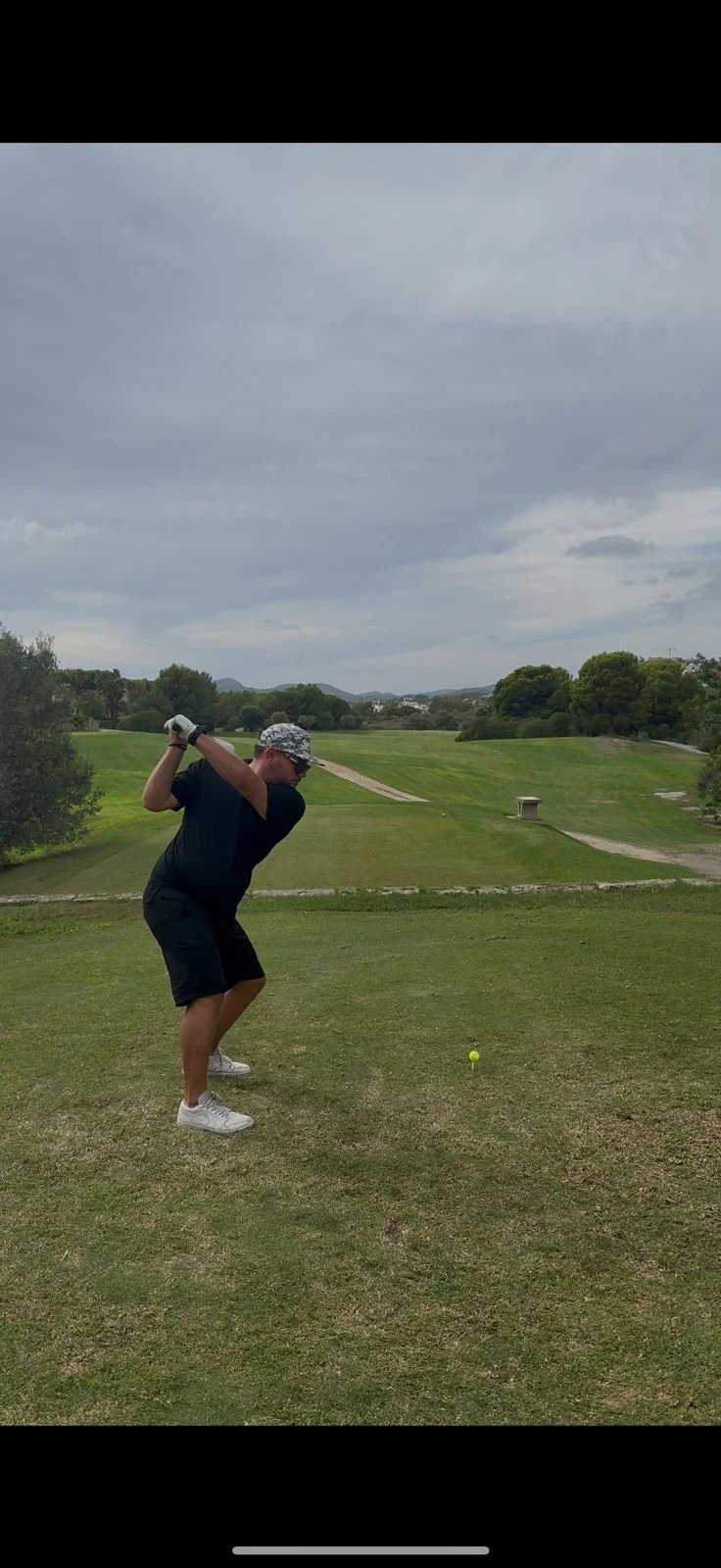 A man in a black T-shirt, shorts, and a hat is swinging a golf club at a golf ball on a tee on a golf course under a cloudy sky.
