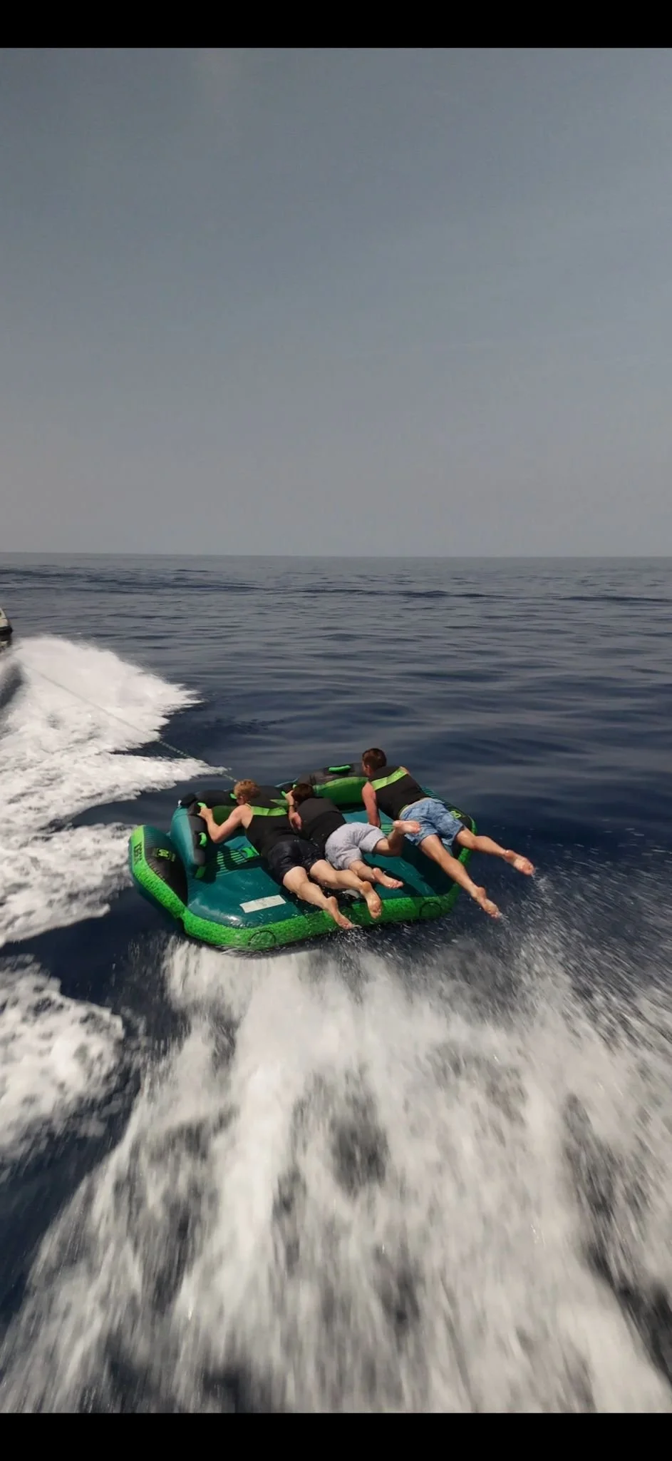 Four people are riding on an inflatable water tube being towed by a boat on open water.